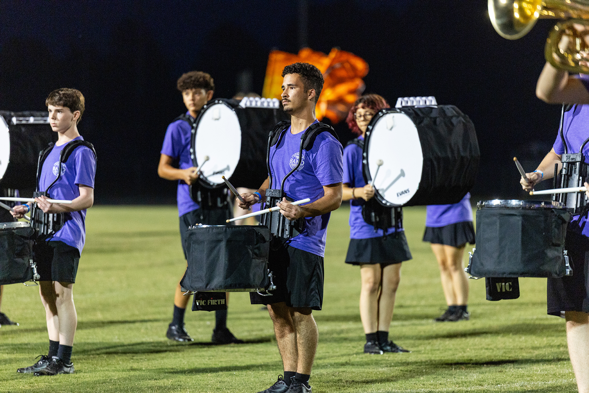 Several individuals in matching uniforms play drums and brass instruments on a green field under a dark sky.