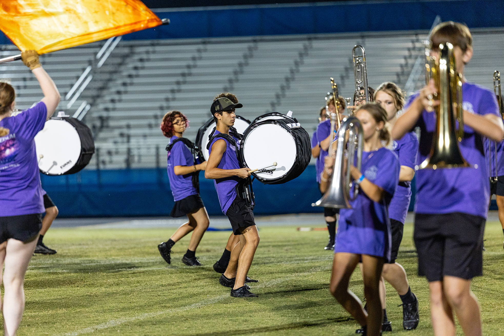 A marching band in purple uniforms performs on a field at dusk, with a flag and instruments.