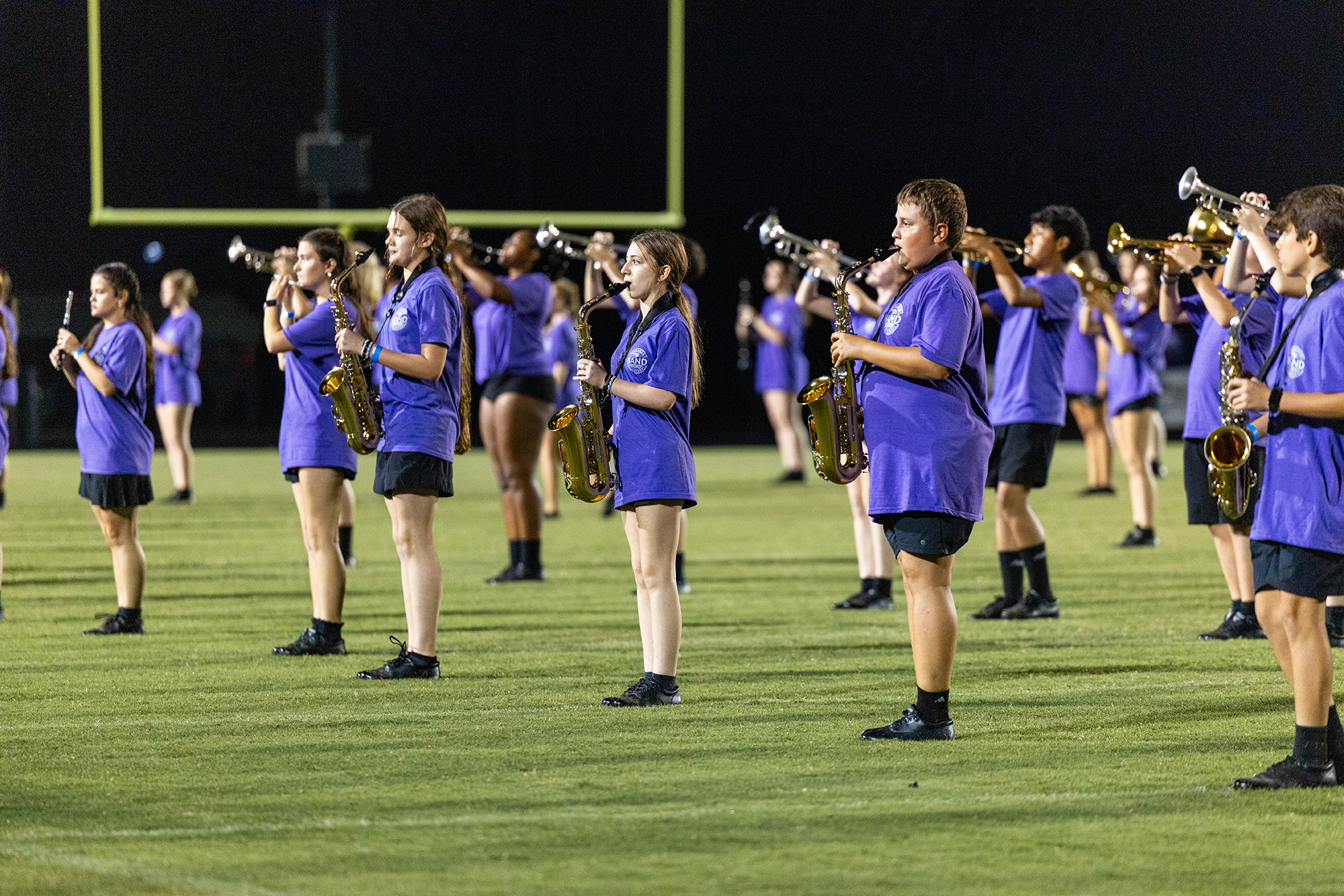 Musicians in purple shirts and black shorts stand on a field holding brass instruments at night.