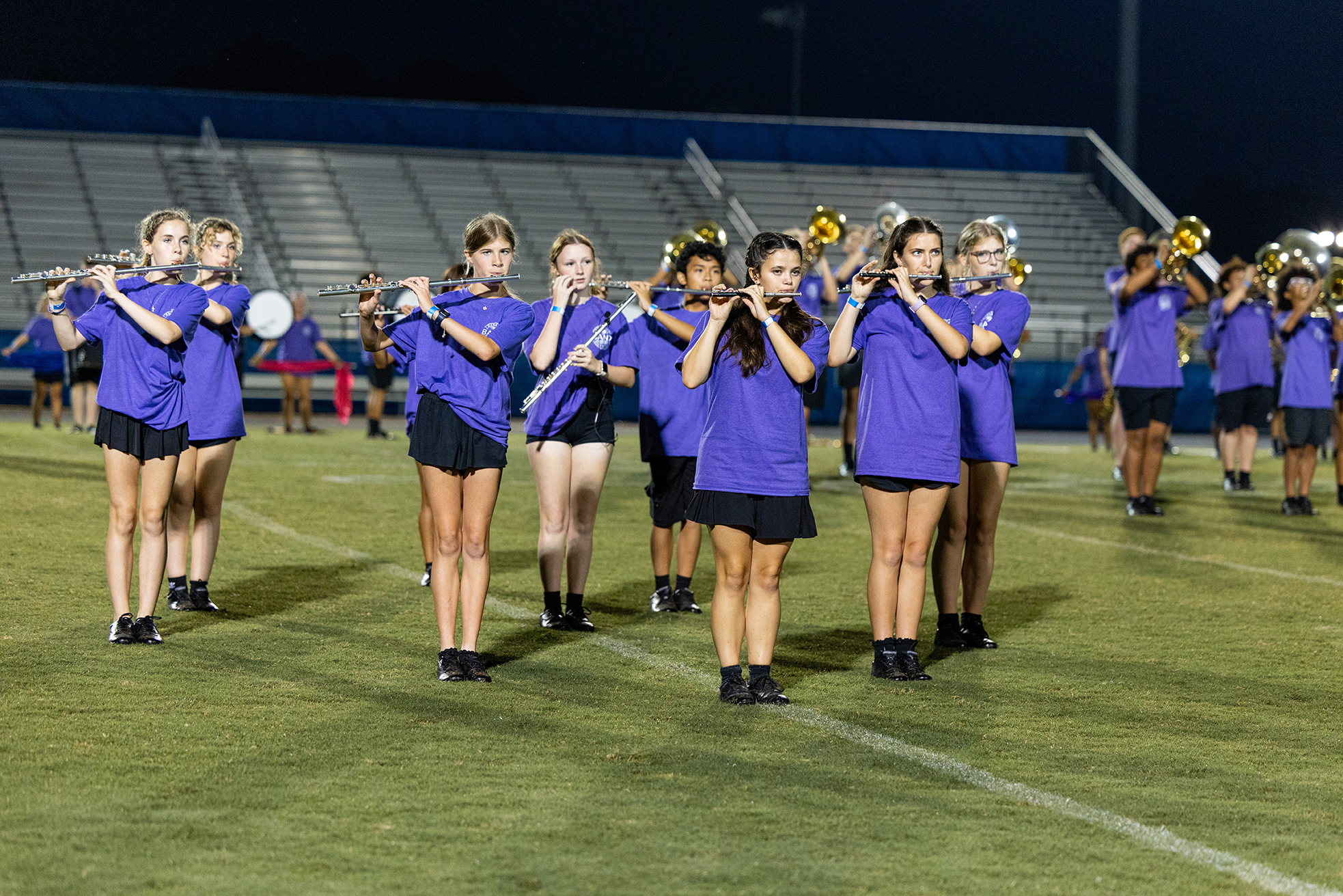 Group of musicians in purple tops and black shorts play instruments on a field at night.
