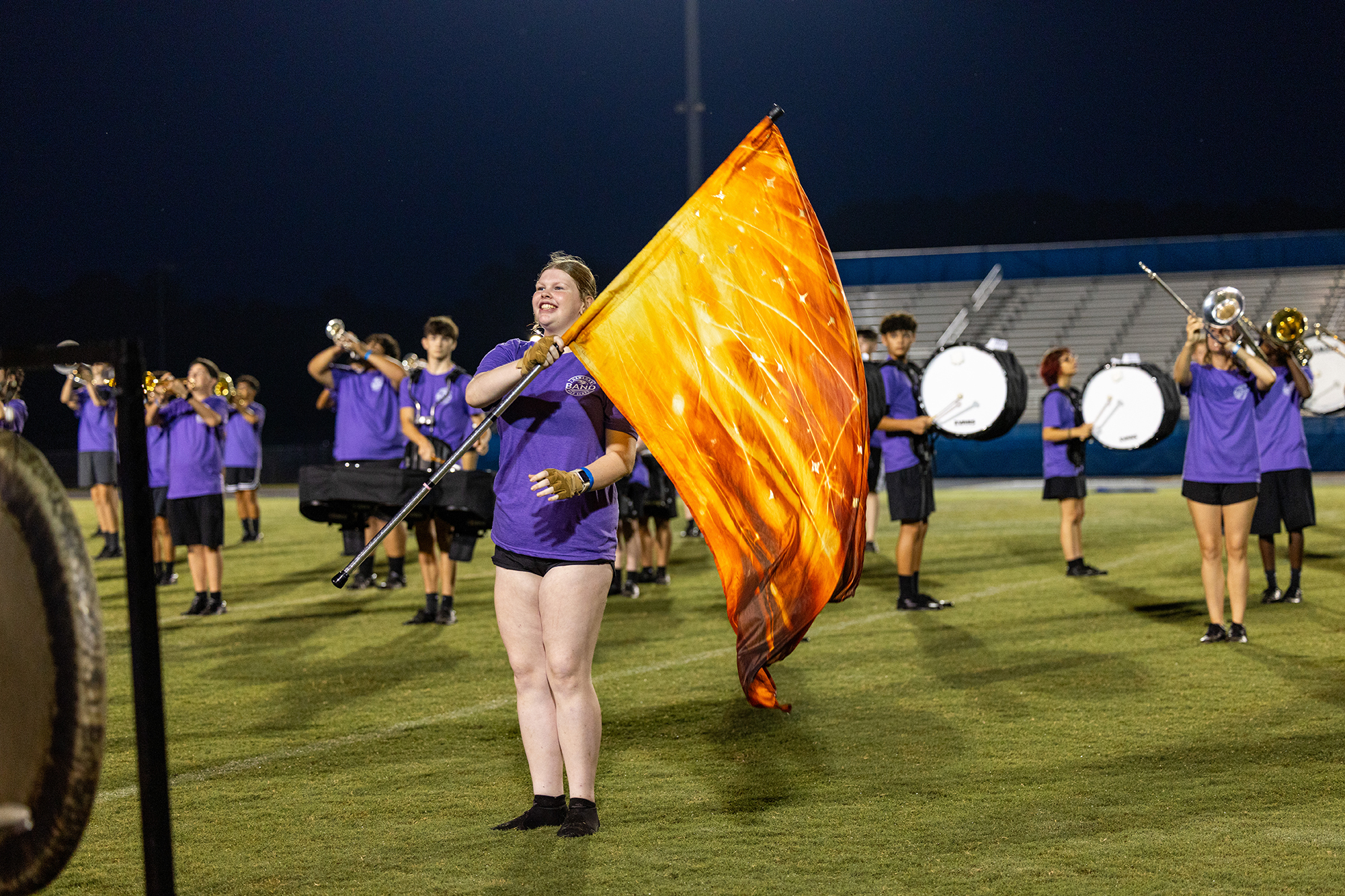 A marching band performs at night; a flag bearer stands front and center, while others play trumpets and drums.