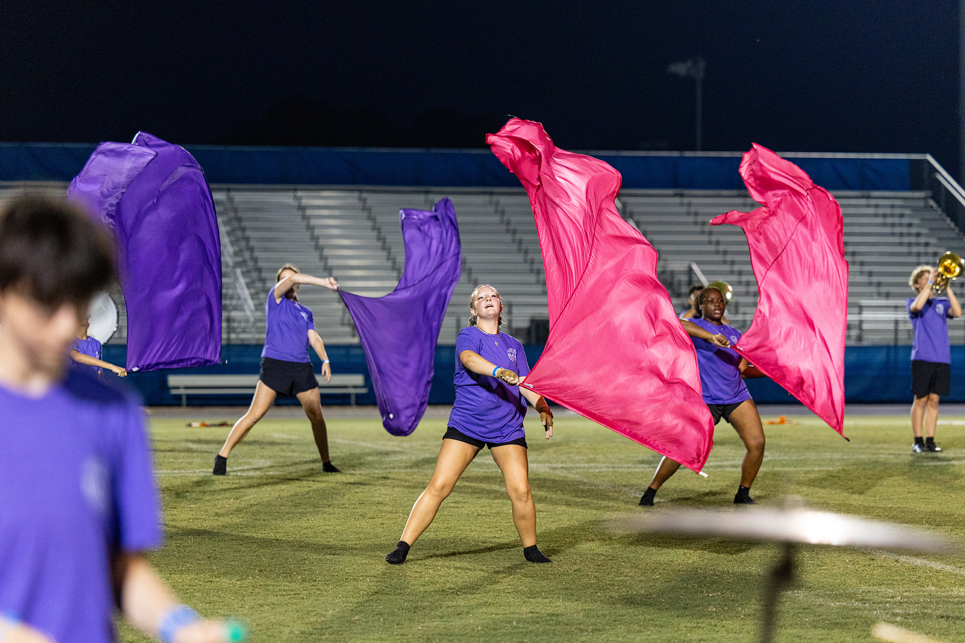 A group of performers on a field with purple and pink fabric, on a field with bleachers.