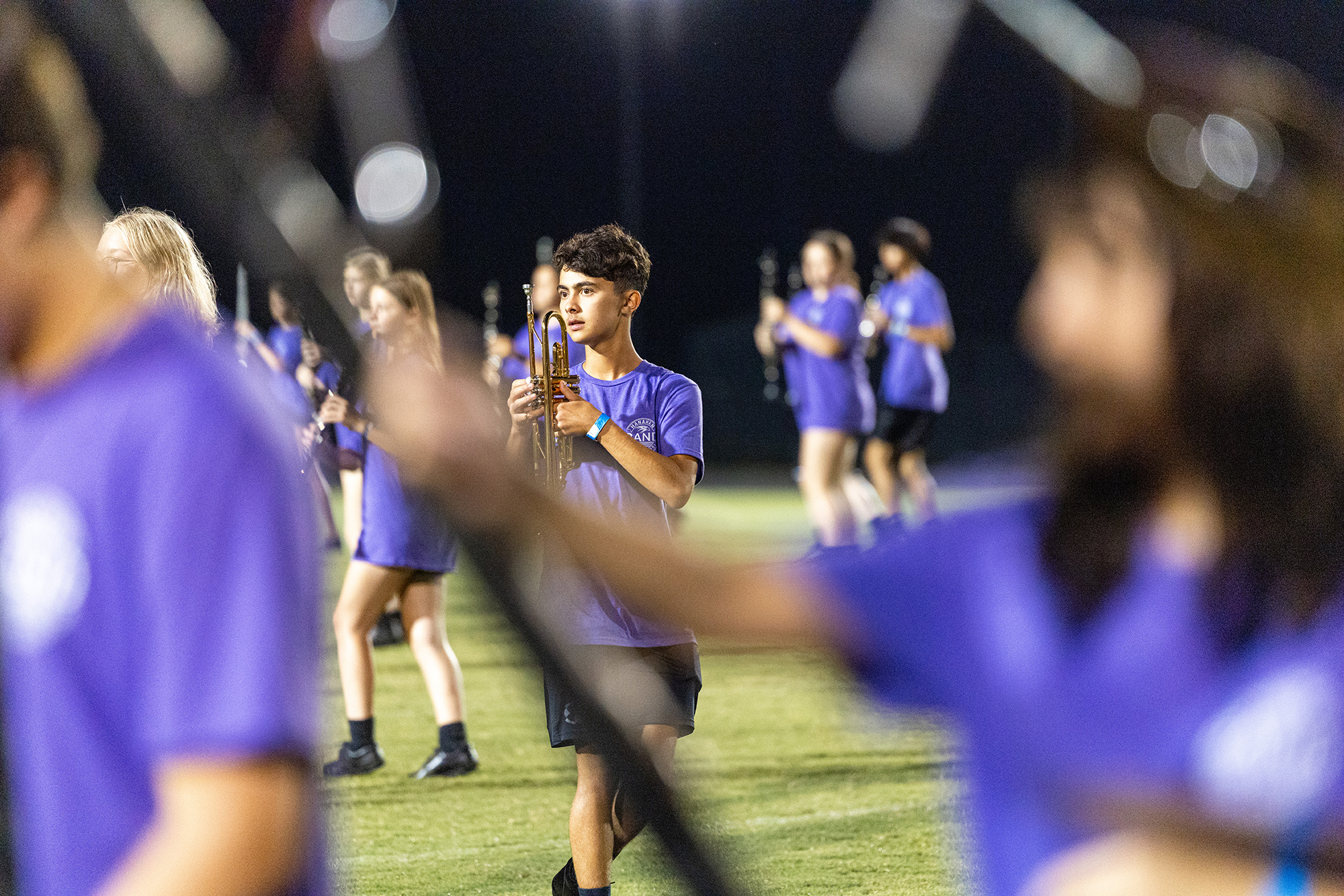A marching band practices on a field at night. Members wear matching purple shirts and black shorts.