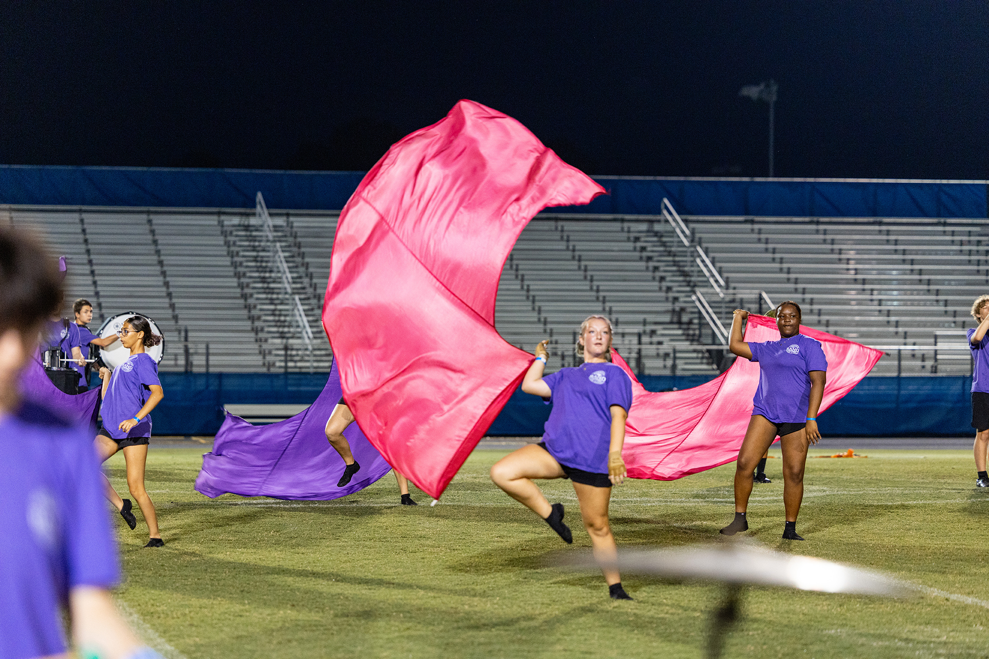 Individuals in purple uniforms perform on a football field, waving pink and purple flags. Empty bleachers are in the background.
