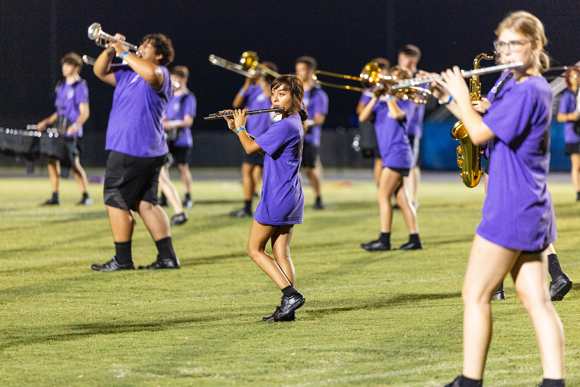 A school band practices on a field at night. Members wear purple uniforms and hold musical instruments.