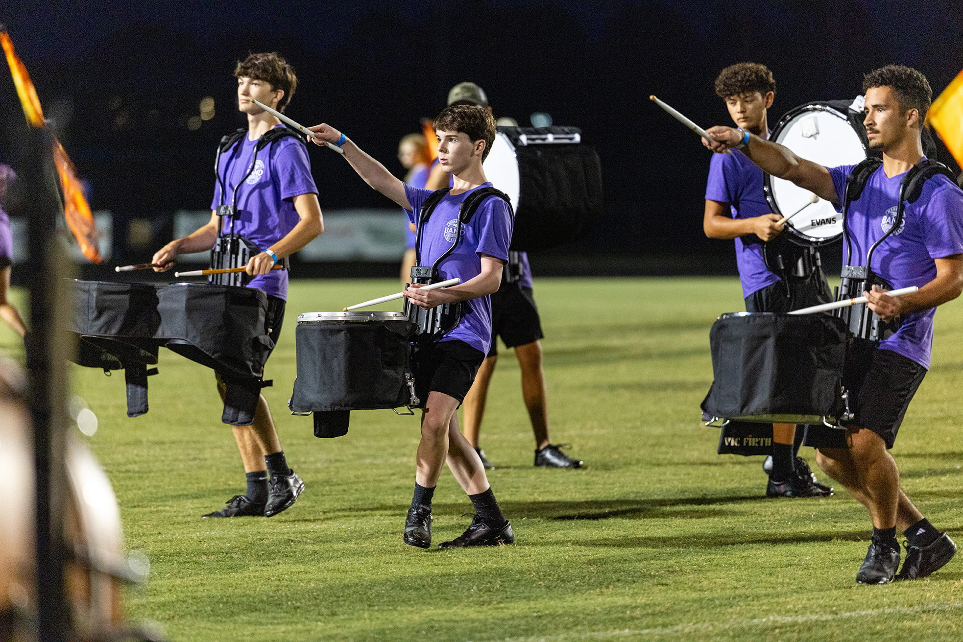 Four drummers with purple shirts and black shorts play drums on a field. Two carry flags.