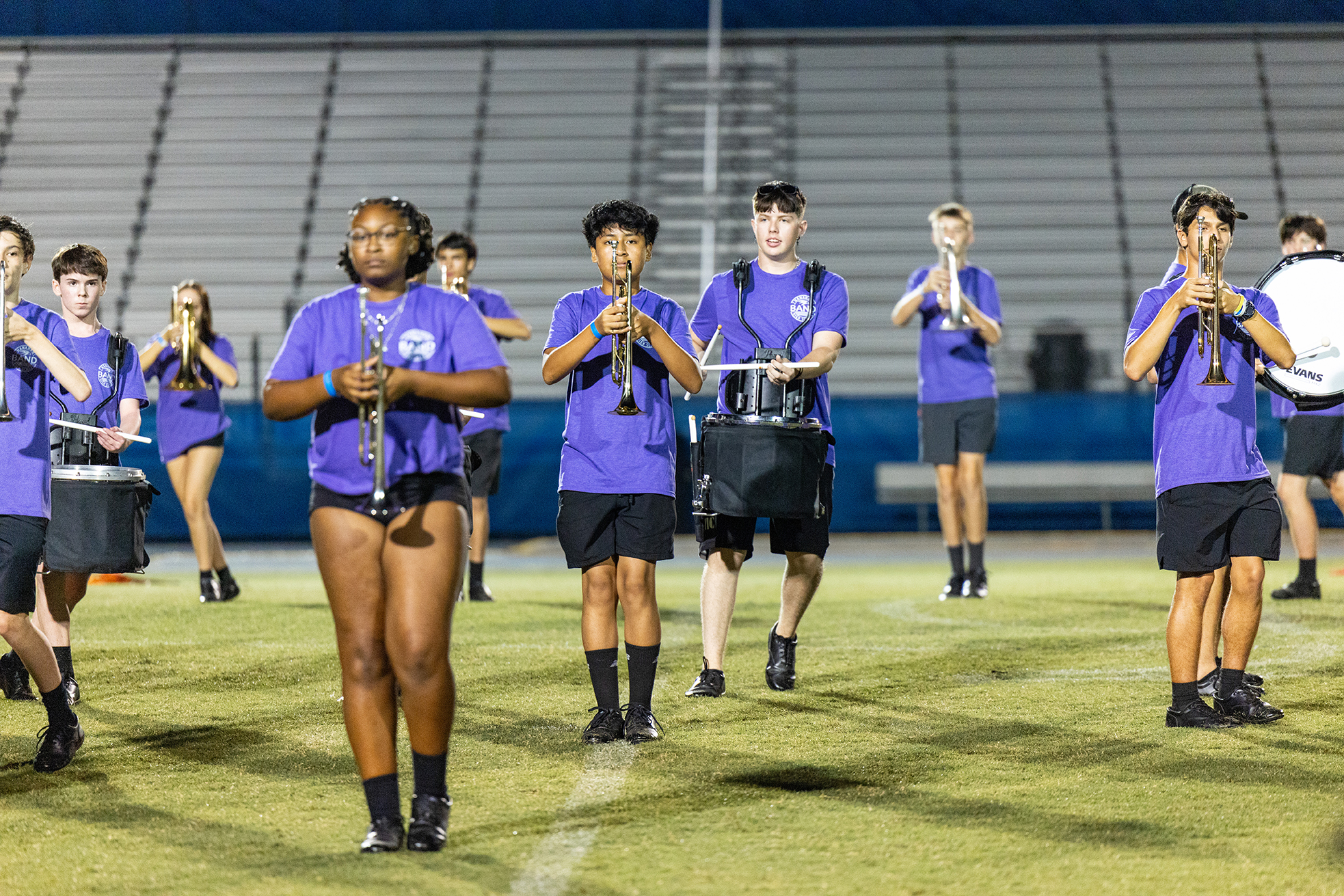 A group of students, all wearing purple shirts and black shorts, playing various instruments on a field.