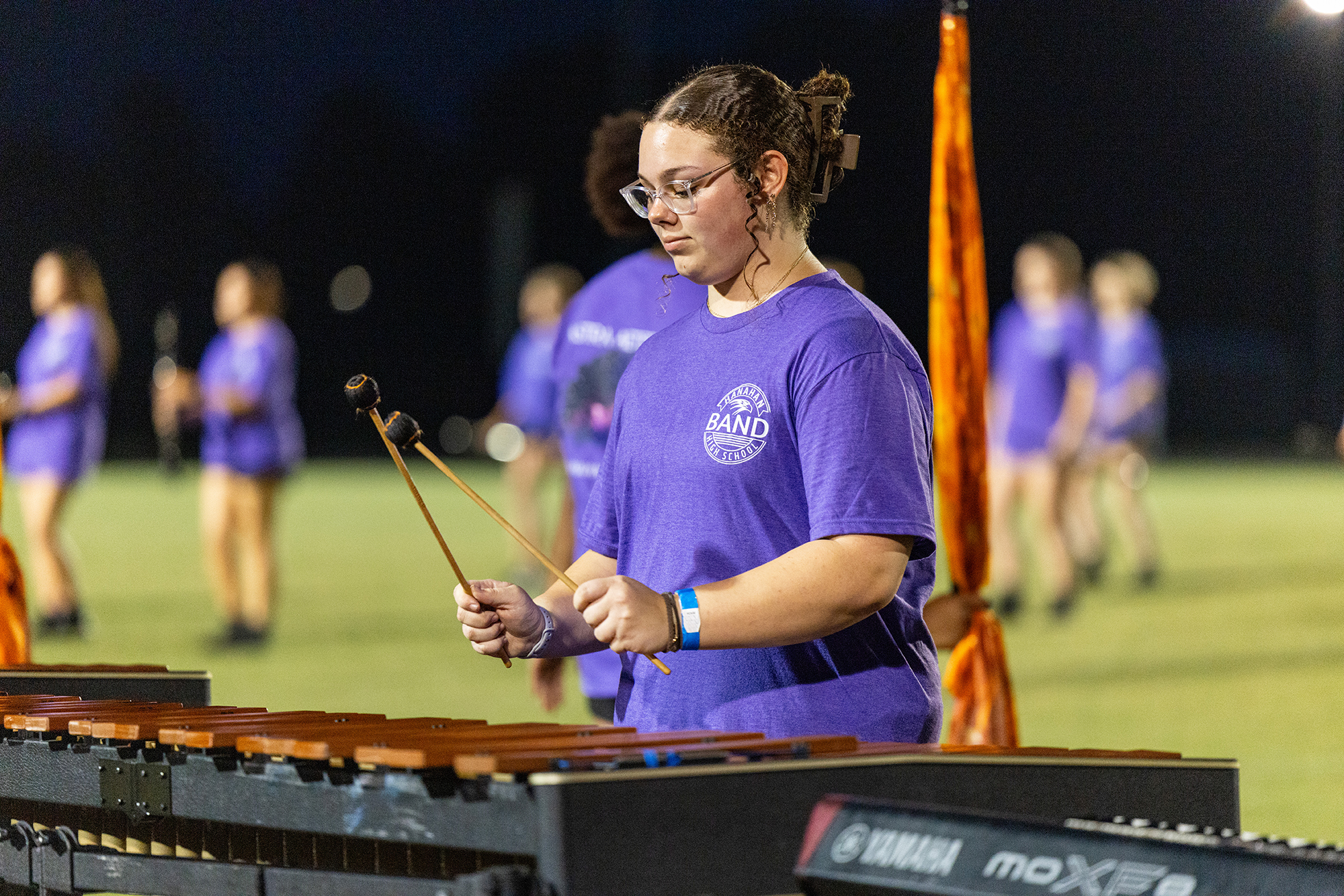 Woman in purple shirt playing xylophone on a field at night with others in purple shirts in background.
