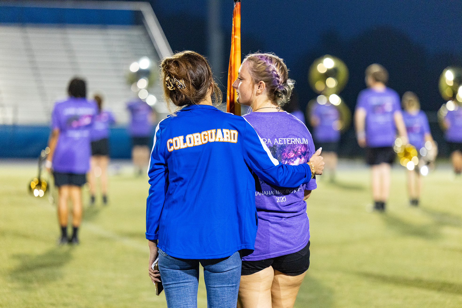 Two women, one in blue, the other in purple, facing each other on a field with a marching band in the background.