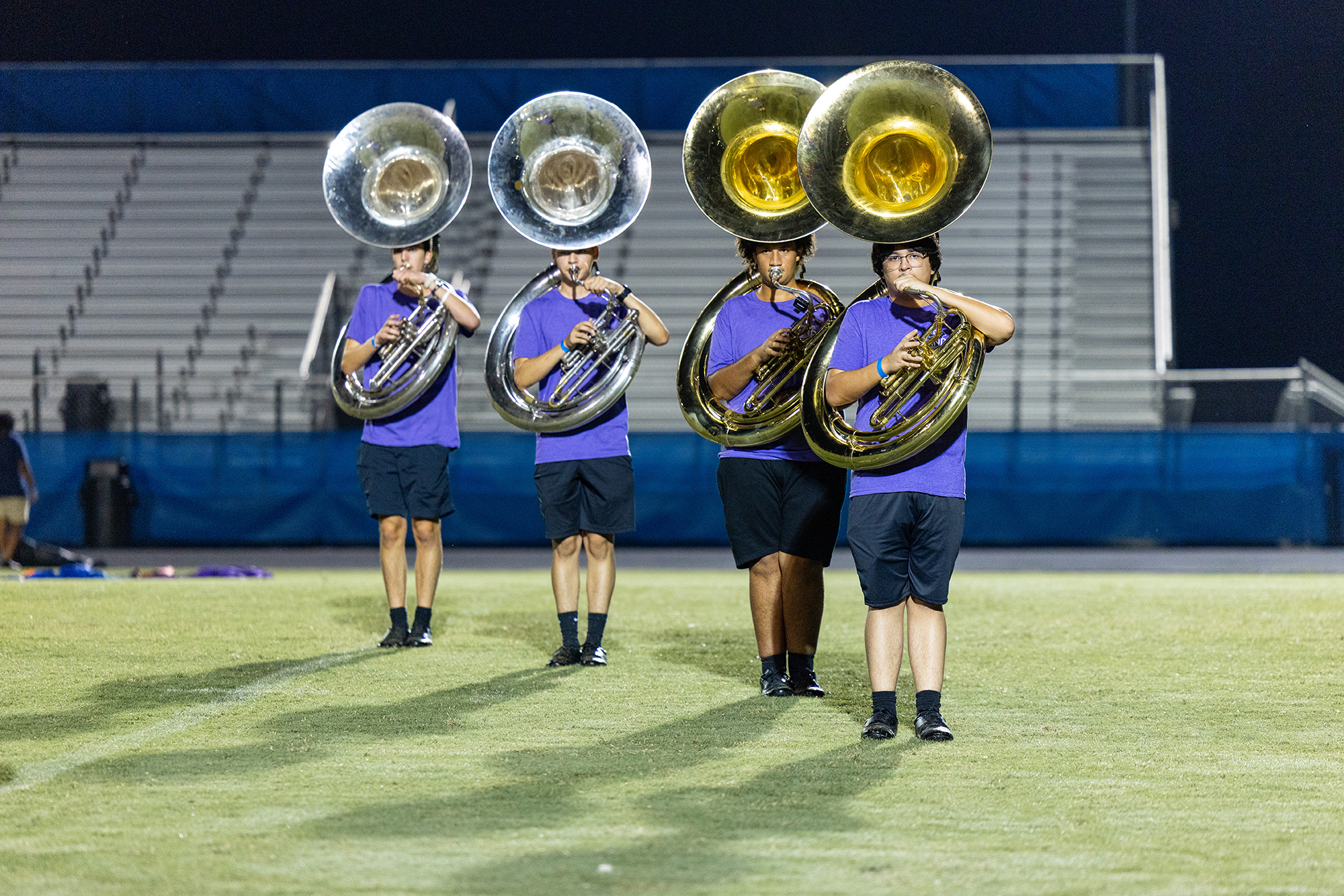 Four musicians play tubas on a football field under blue lights. They wear matching uniforms.