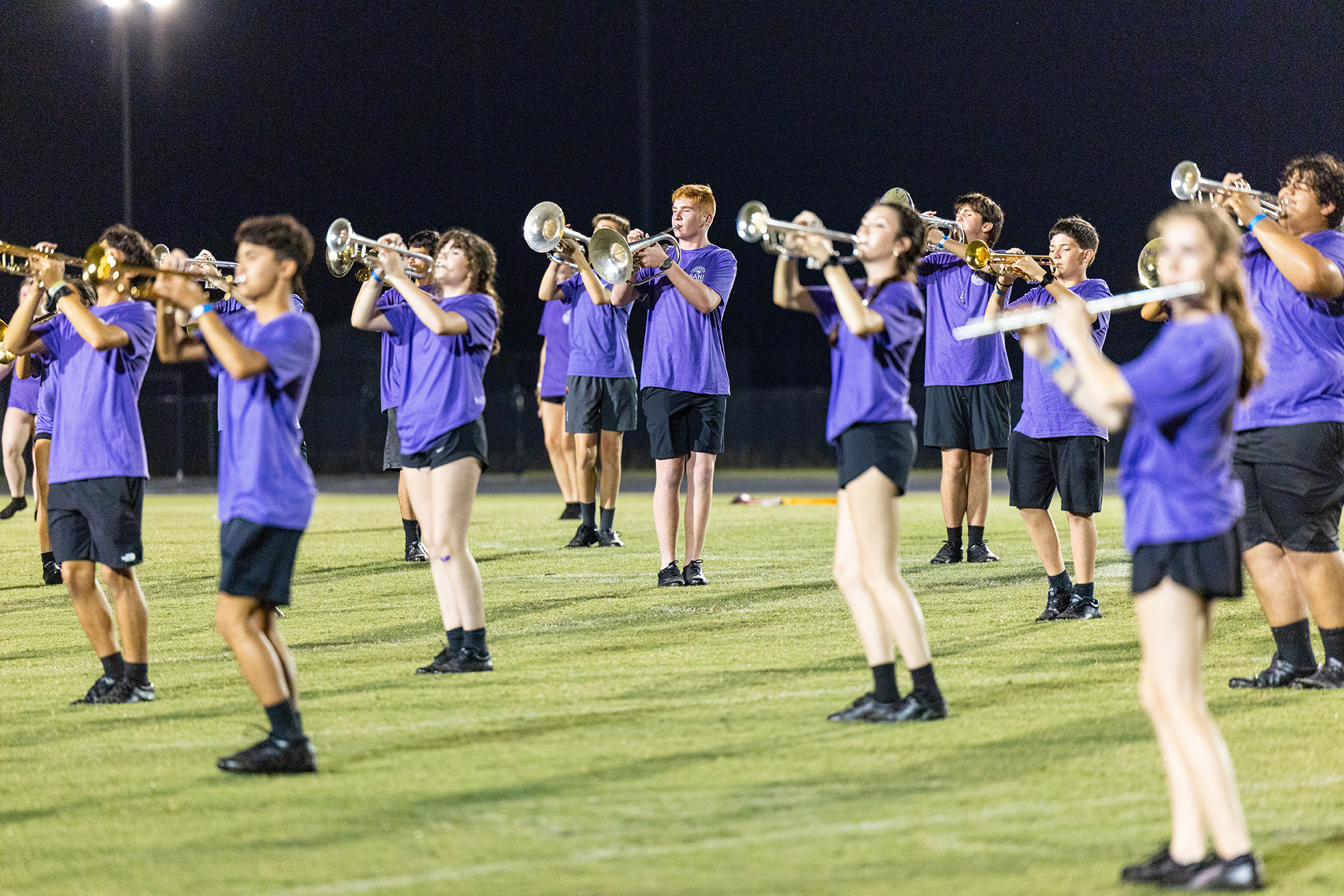 A group of musicians in purple shirts and black shorts play brass and flute instruments on a field.