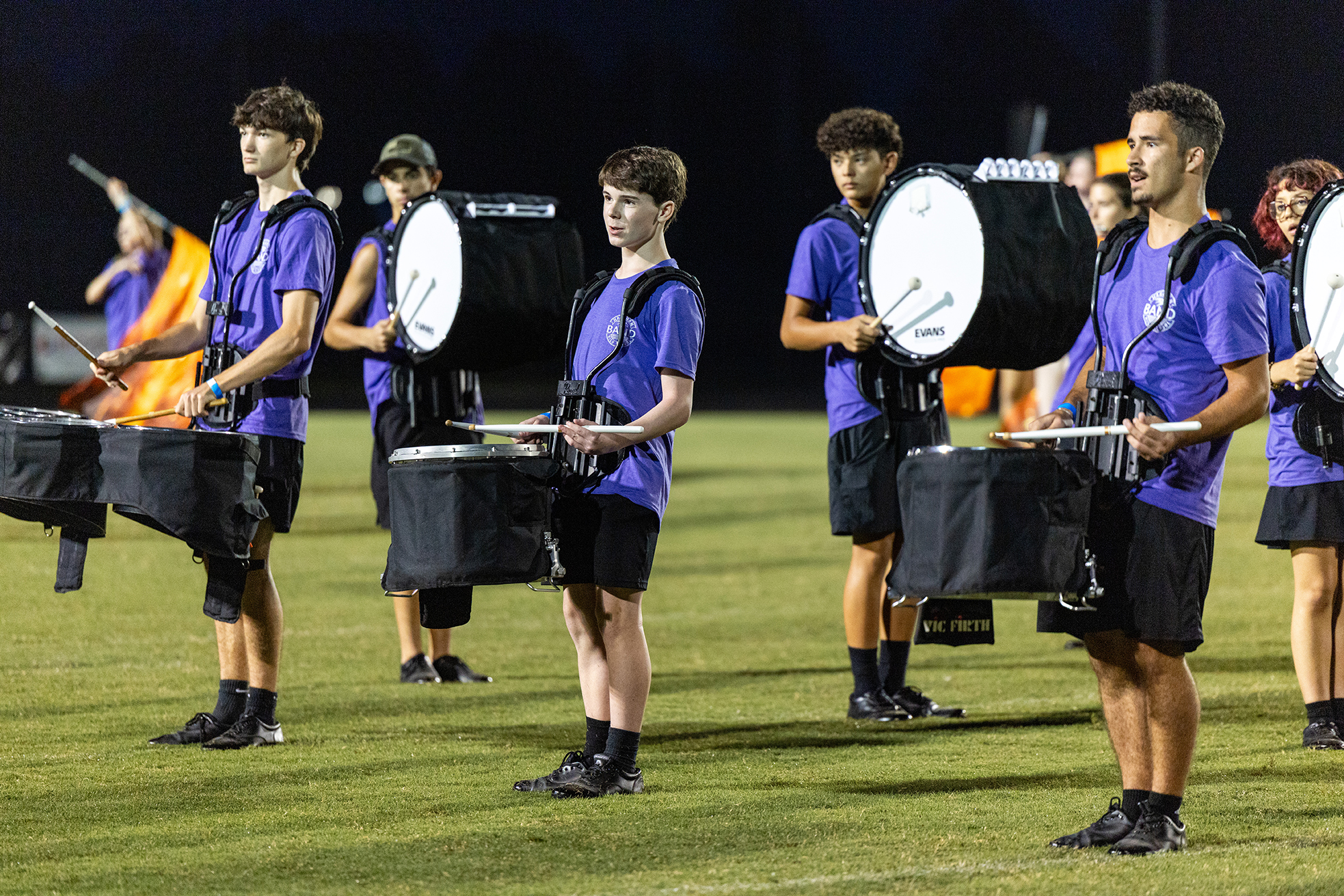 Six individuals in purple uniforms and black shorts, holding drumsticks and drums, lined up on a field.