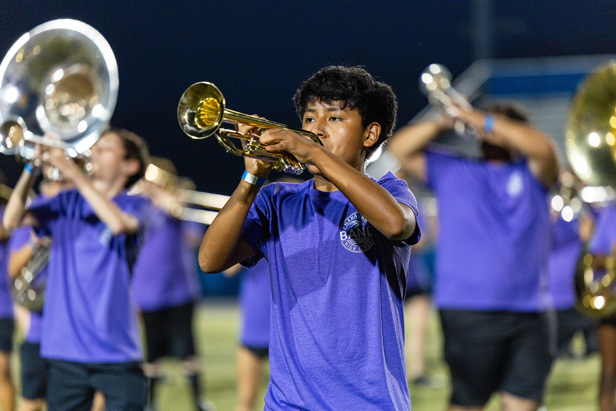 A group of musicians in purple shirts and black shorts, playing trumpets and tubas on a field.