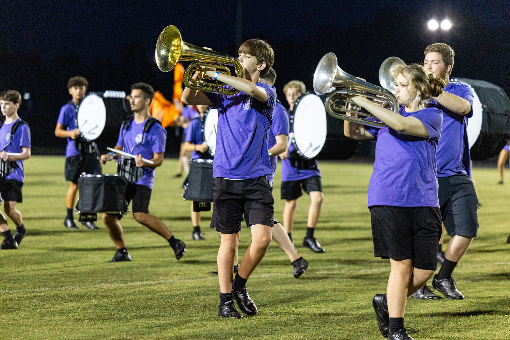 A band on a field, each wearing purple shirts, black shorts, and black shoes, playing instruments.