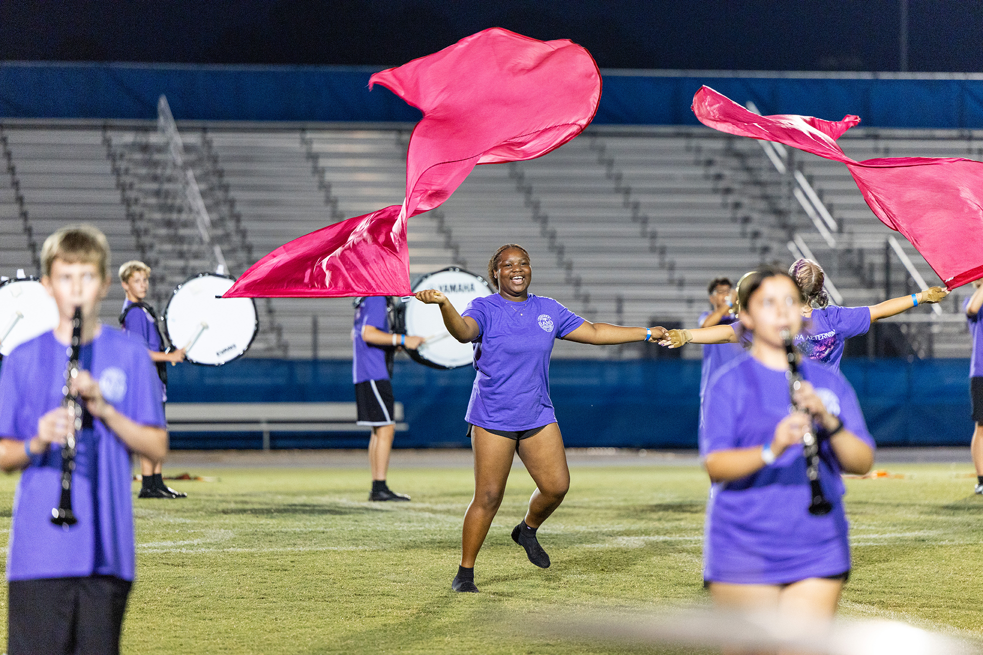 Members of a marching band with pink flags performing on a field in front of empty bleachers.