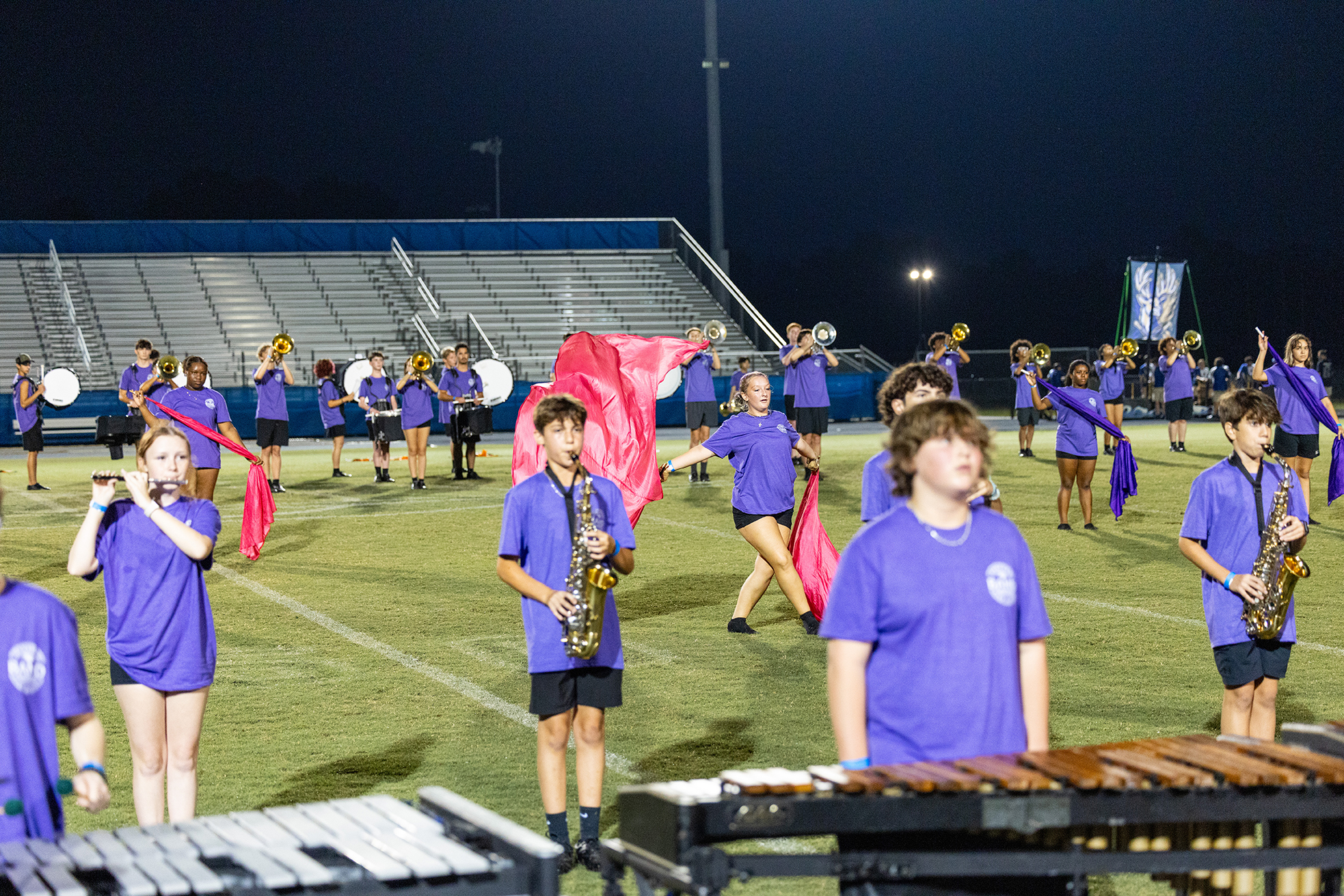 A marching band in purple uniforms performs on a field with various instruments like saxophones and xylophones.