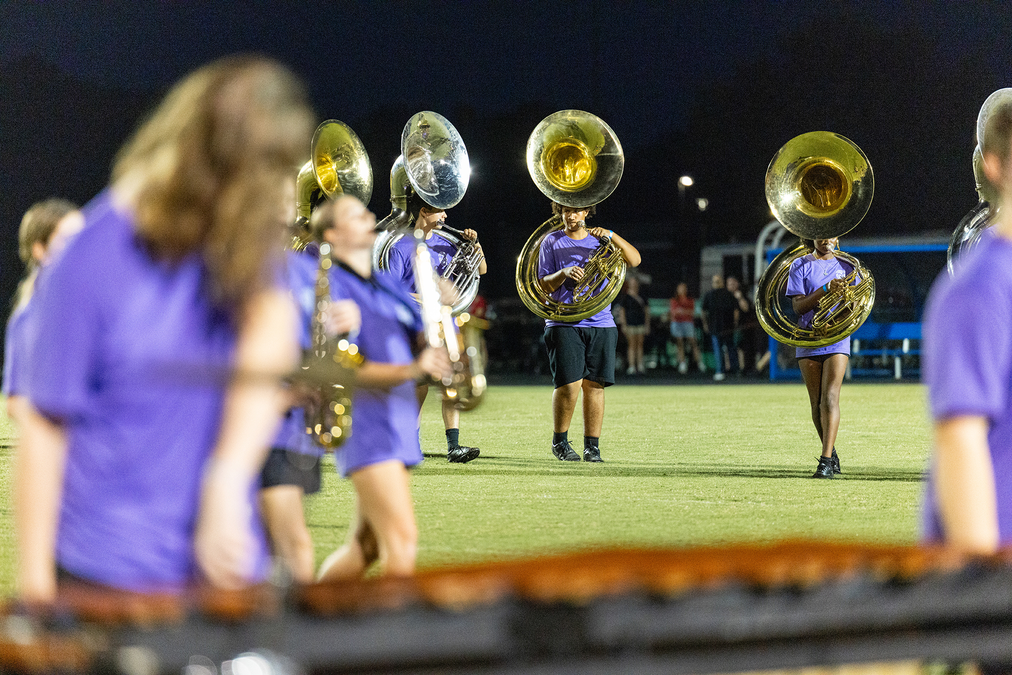 A group of musicians wearing purple uniforms play brass instruments on a field at night.