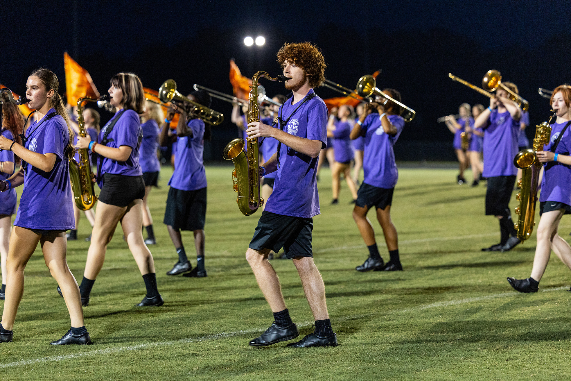 A marching band with members in blue uniforms performs on a field at night, holding instruments like trombones, saxophones, and trumpets.