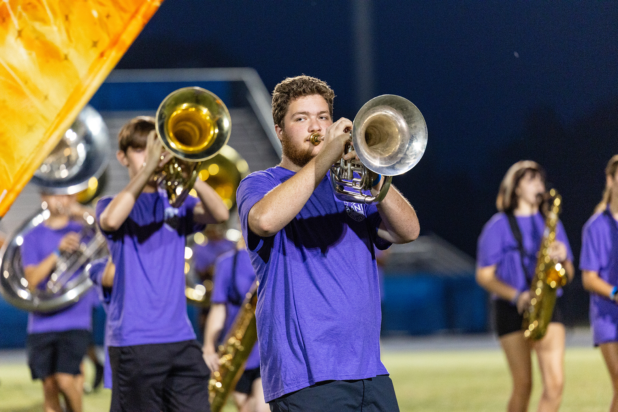 A group of musicians in purple shirts play trumpets and tubas on a field at night.
