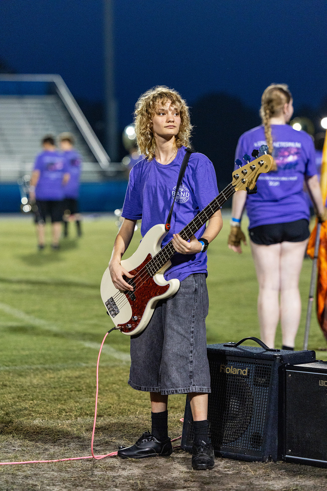 A person with curly hair holds a guitar on a football field. Another person stands behind.