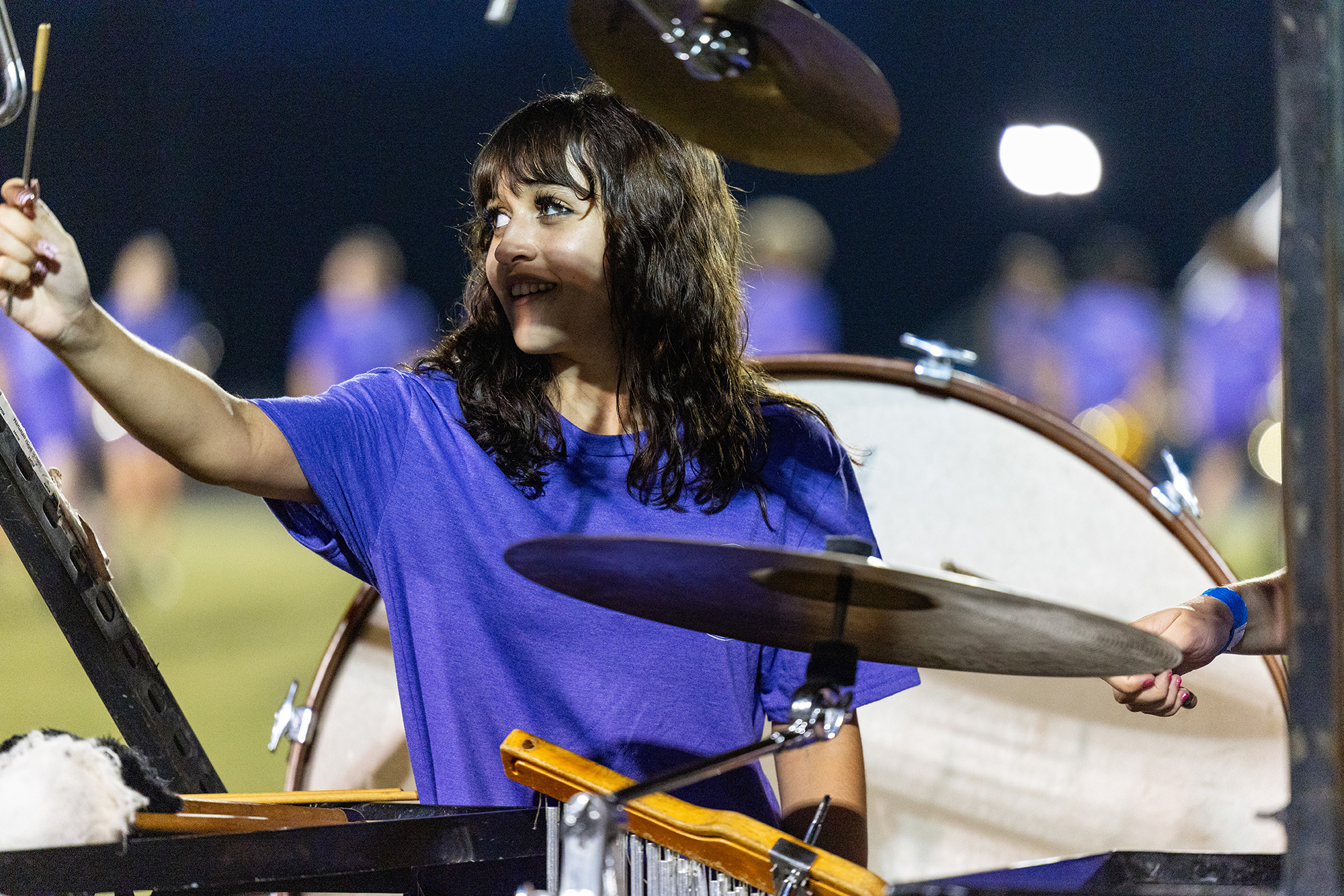 A woman in a blue shirt plays the xylophone outdoors, with a drum set behind her and blurred teammates.