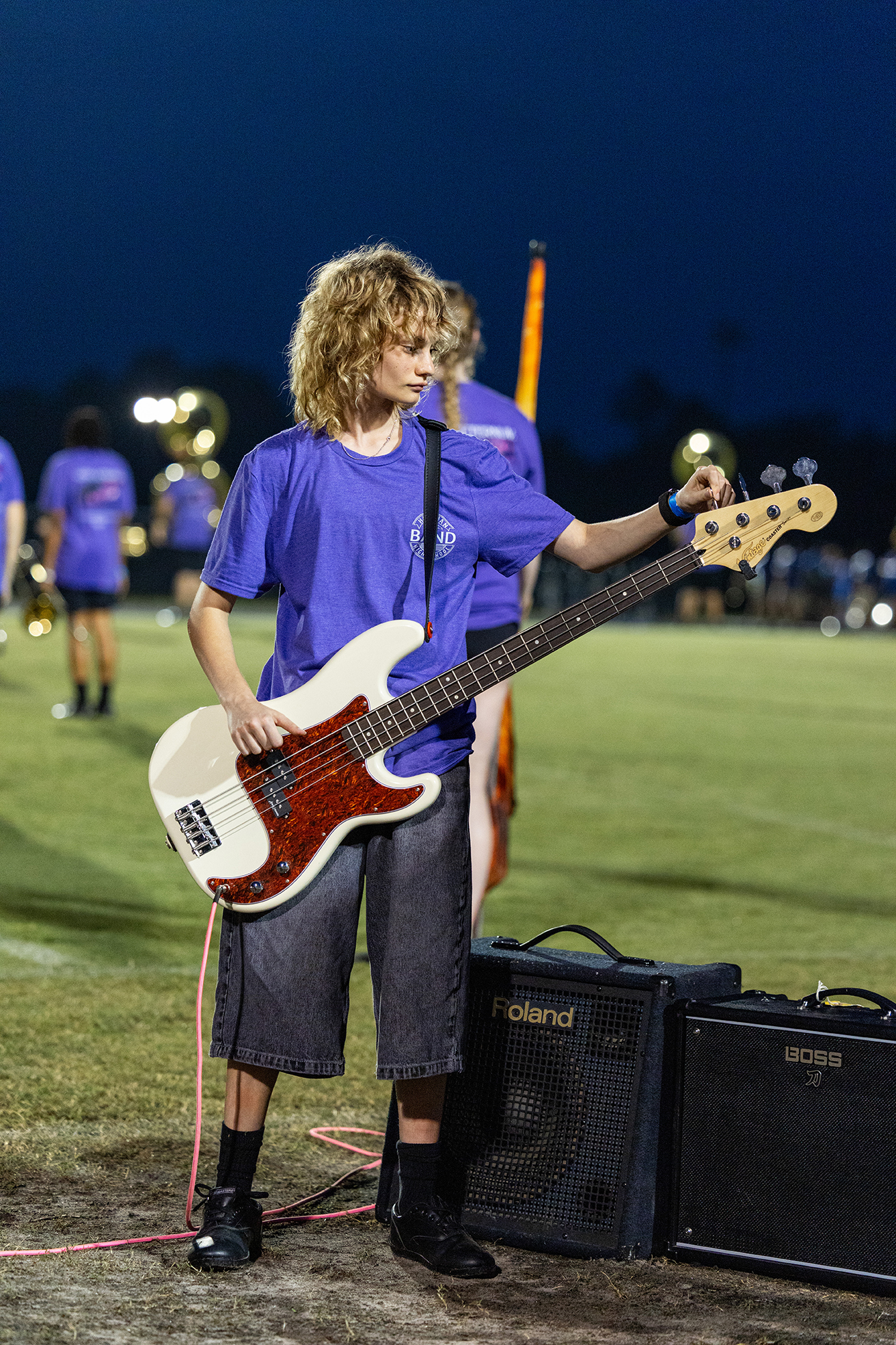 A person in a blue shirt plays a white bass guitar on a field with others in the background.