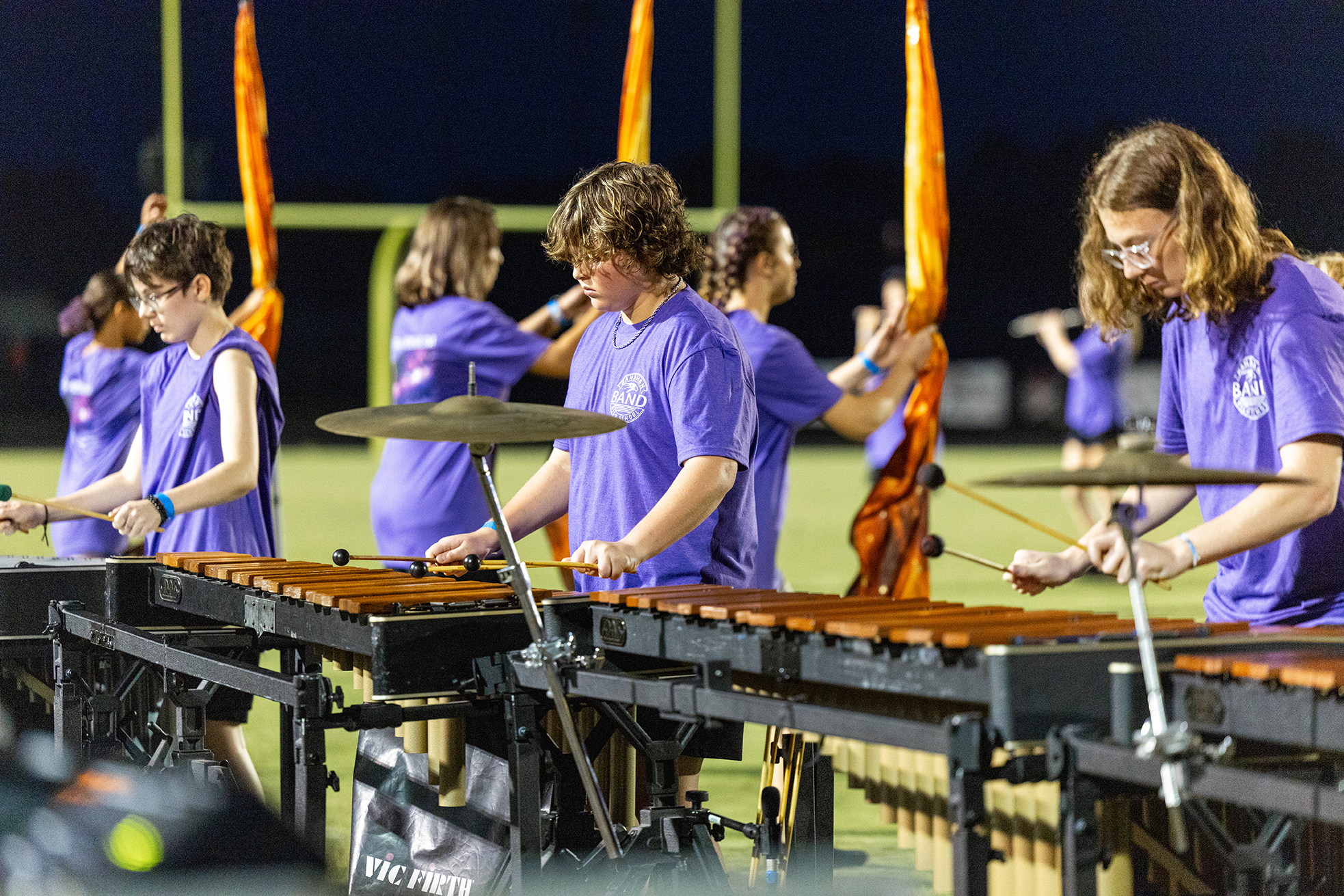 Seven people wearing purple shirts and glasses playing xylophones on a football field at night.