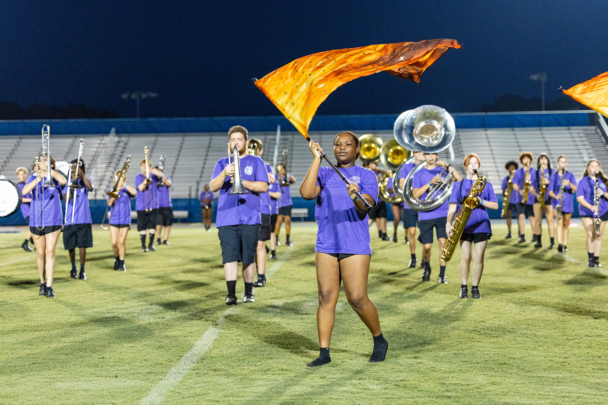 A marching band performs on a field with orange flags, under stadium lights at night.