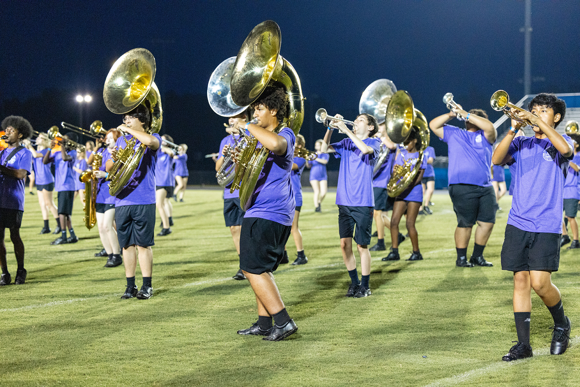 A group of musicians in blue shirts and black shorts plays brass instruments on a field.
