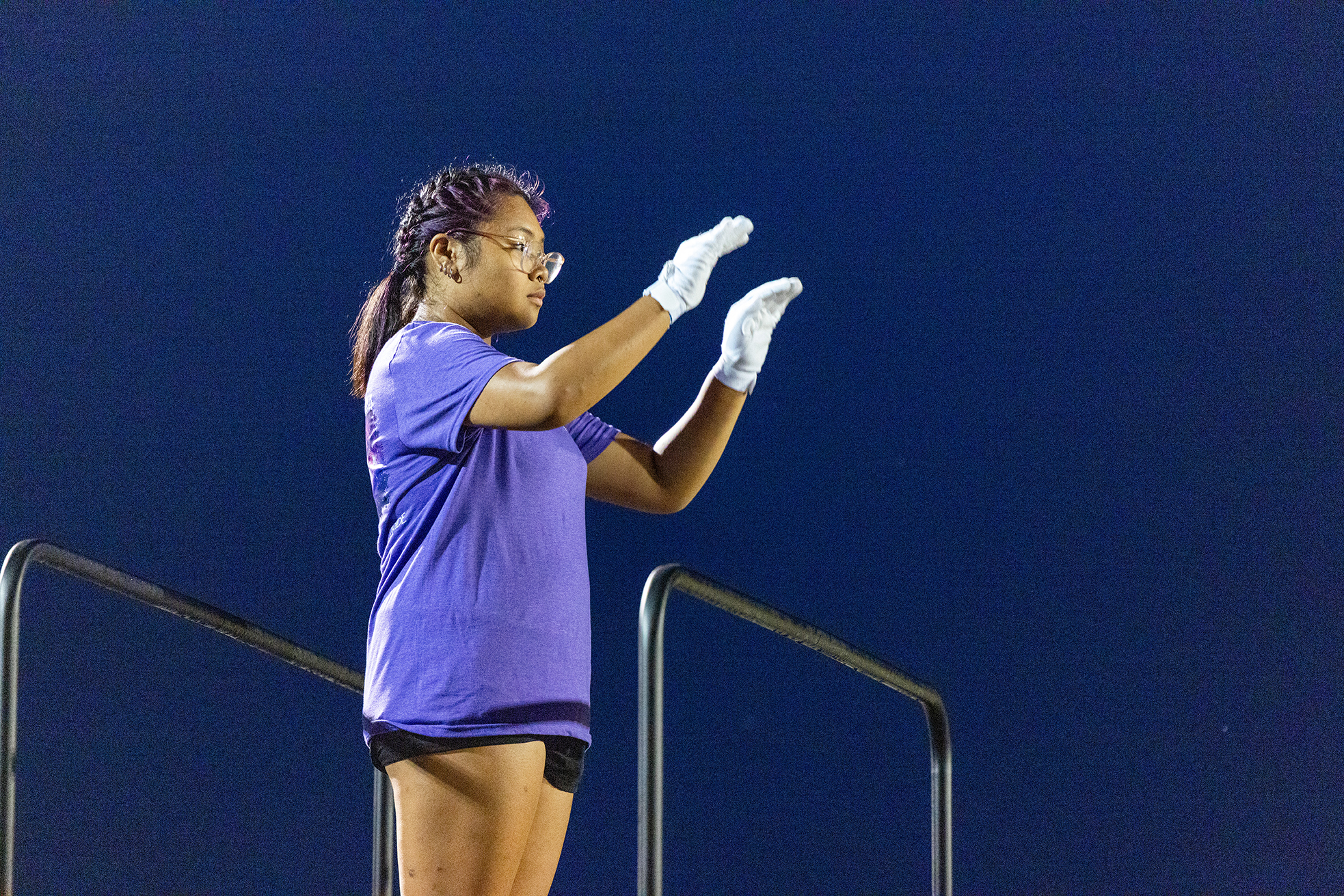 A cheerleader in a purple shirt and white gloves stands on bleachers with arms raised.