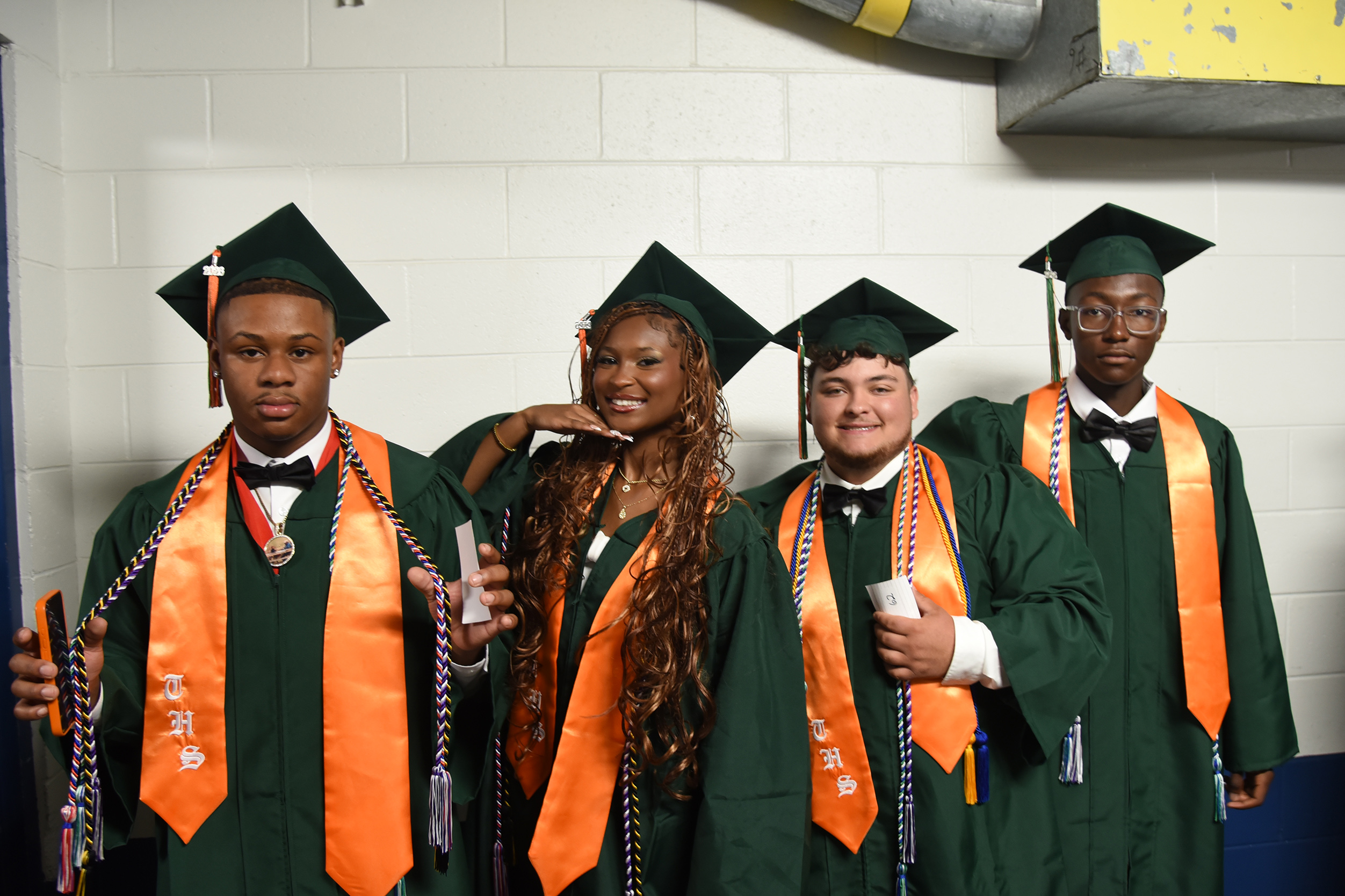 Four people in green caps and gowns pose for a photo. Each wears orange sashes with white tassels.