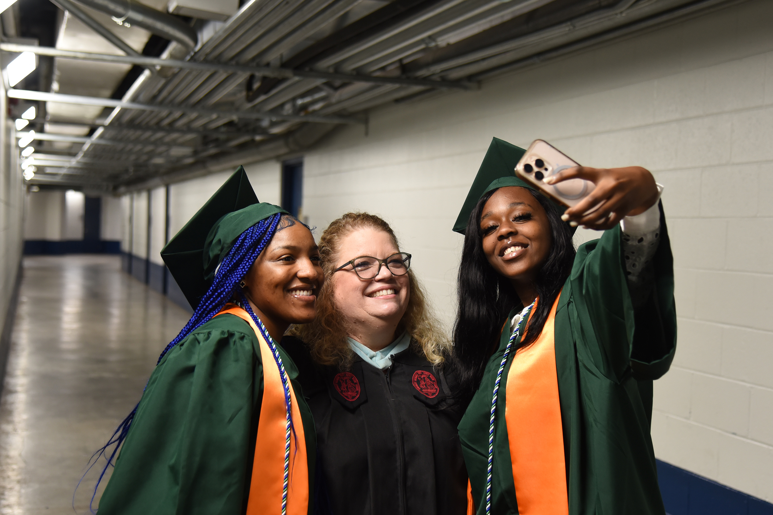 Three women in graduation gowns stand in a tunnel. Two are smiling and one holds a phone.