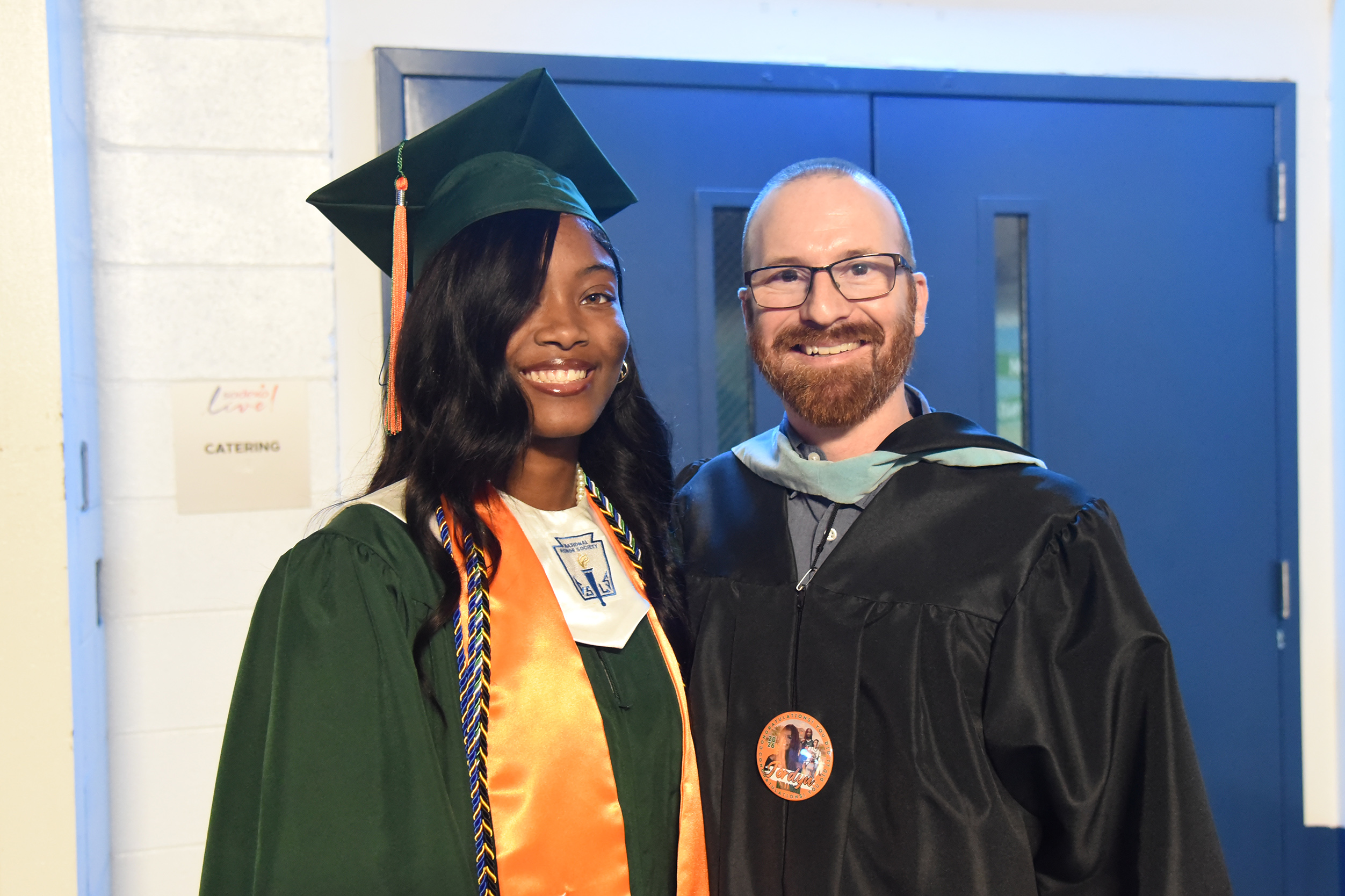 A woman and man, both in graduation attire, smiling in front of a blue door.