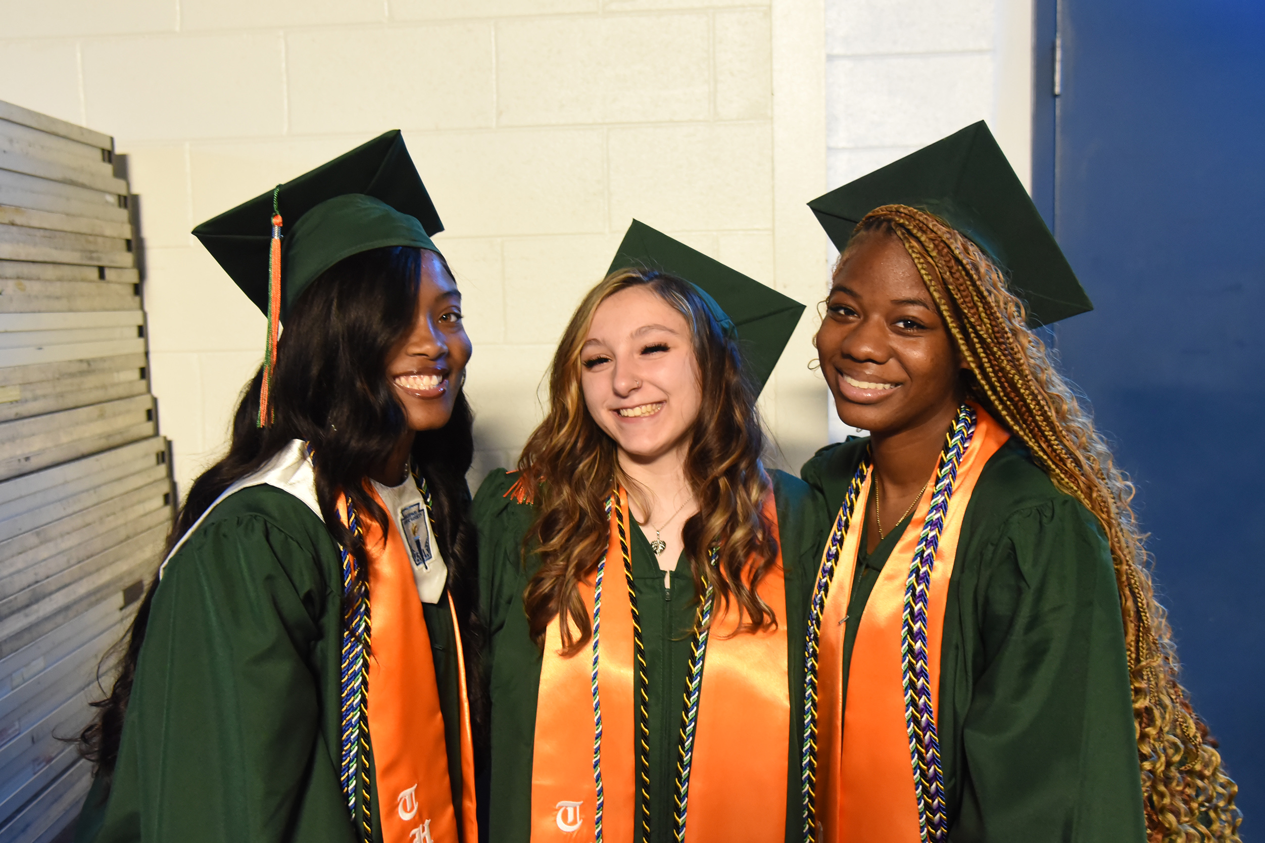 Three women in green gowns and caps, smiling. One wears an orange sash, another a blue and yellow sash.