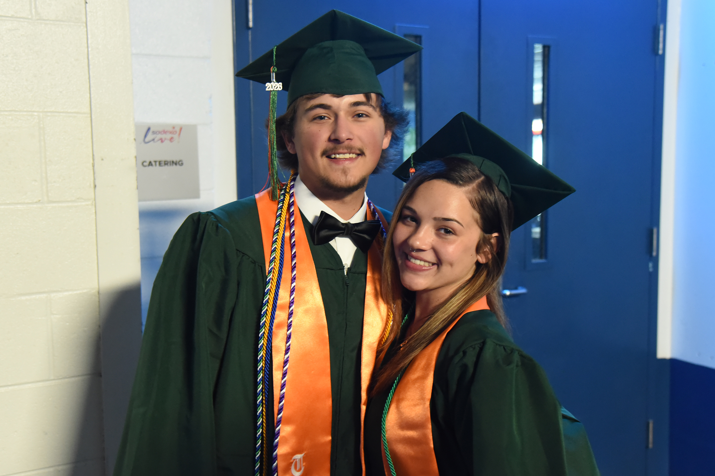 A man and a woman in green graduation gowns and caps stand next to a blue door.