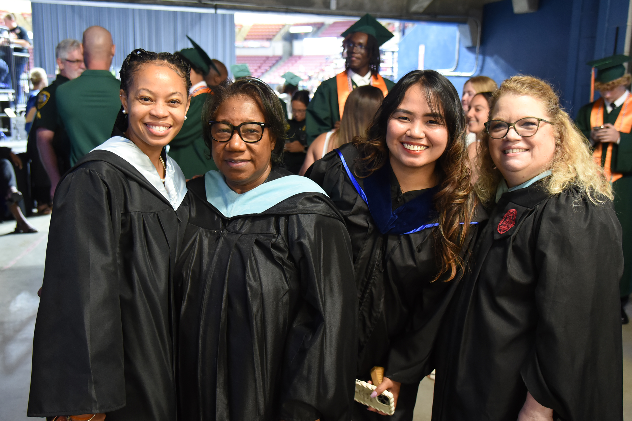 Four women in black gowns smile for a photo at an outdoor event, likely a graduation.