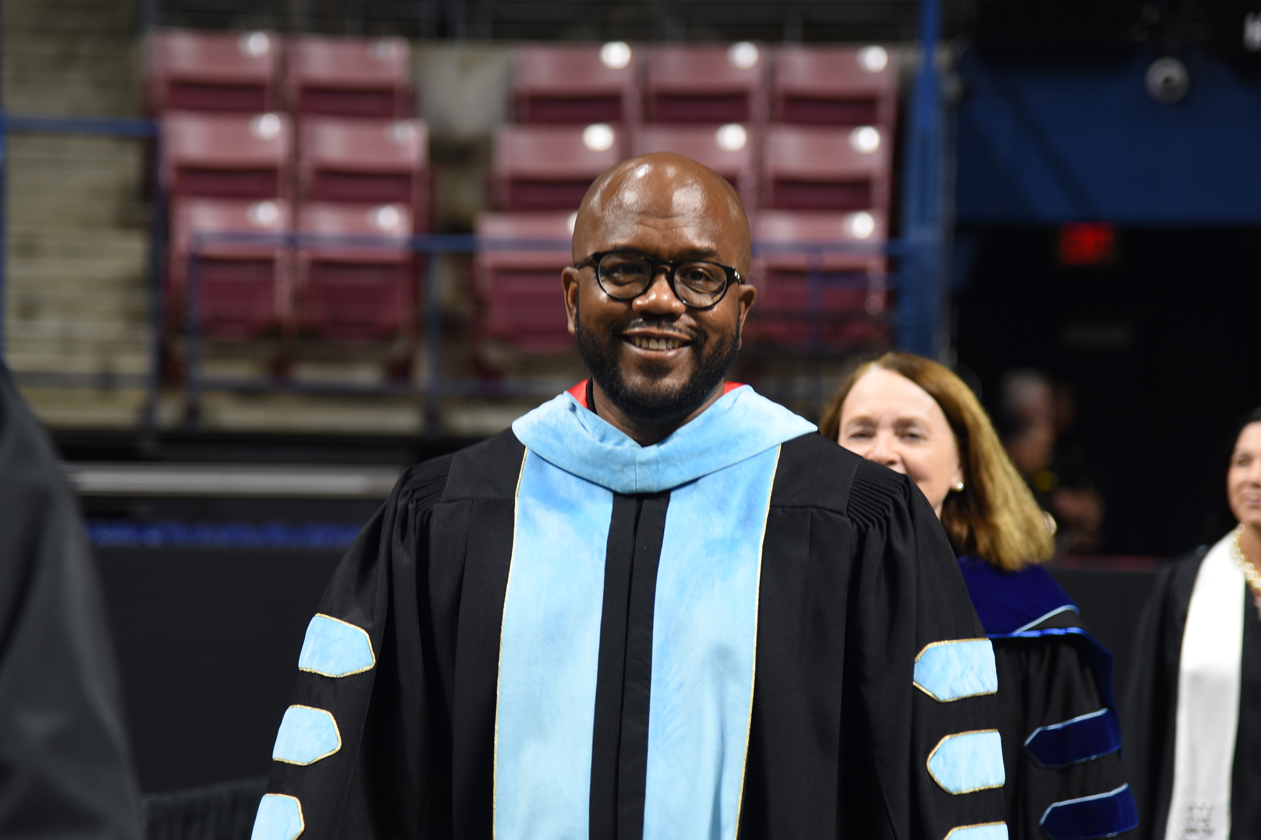 A man in academic regalia smiles, standing in front of rows of empty red chairs and a blurred figure.