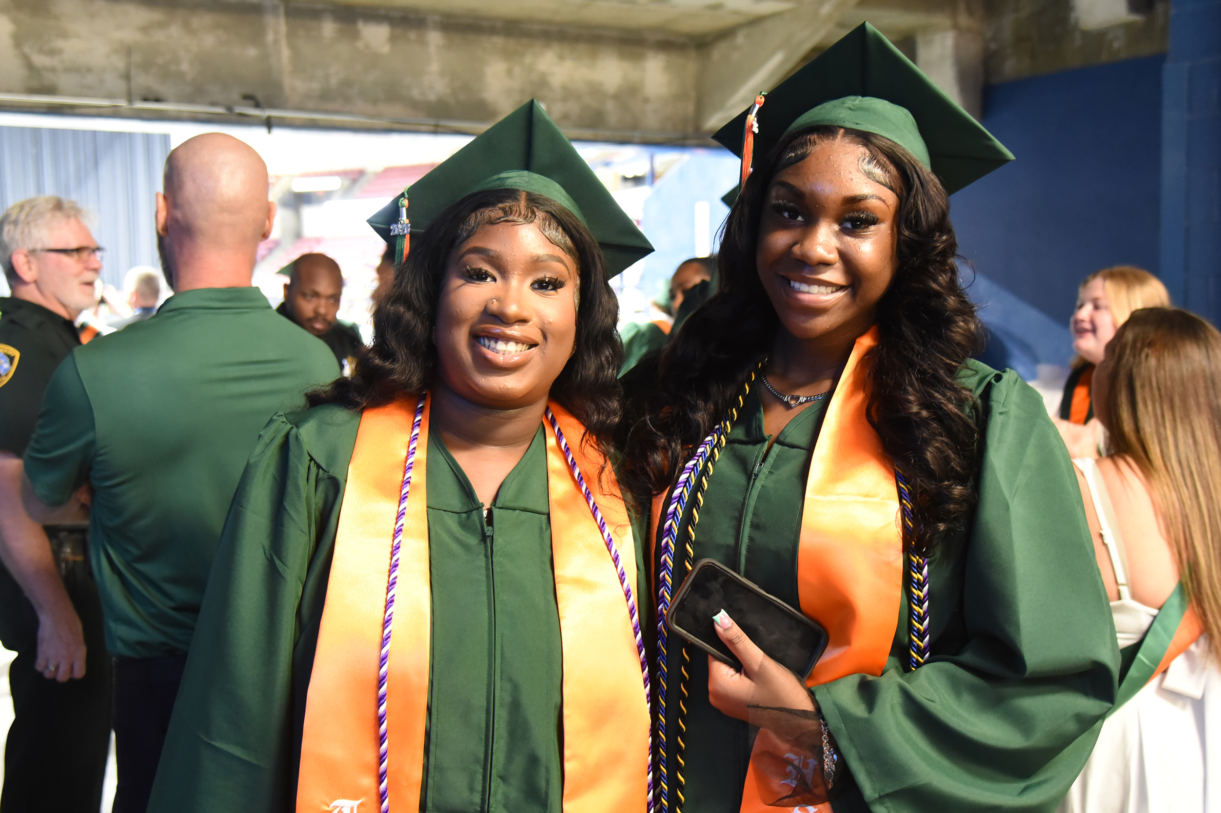 Two women in green gowns and orange stoles smile, one holding a phone. Behind them, a man in a green shirt stands with two others, one in glasses.