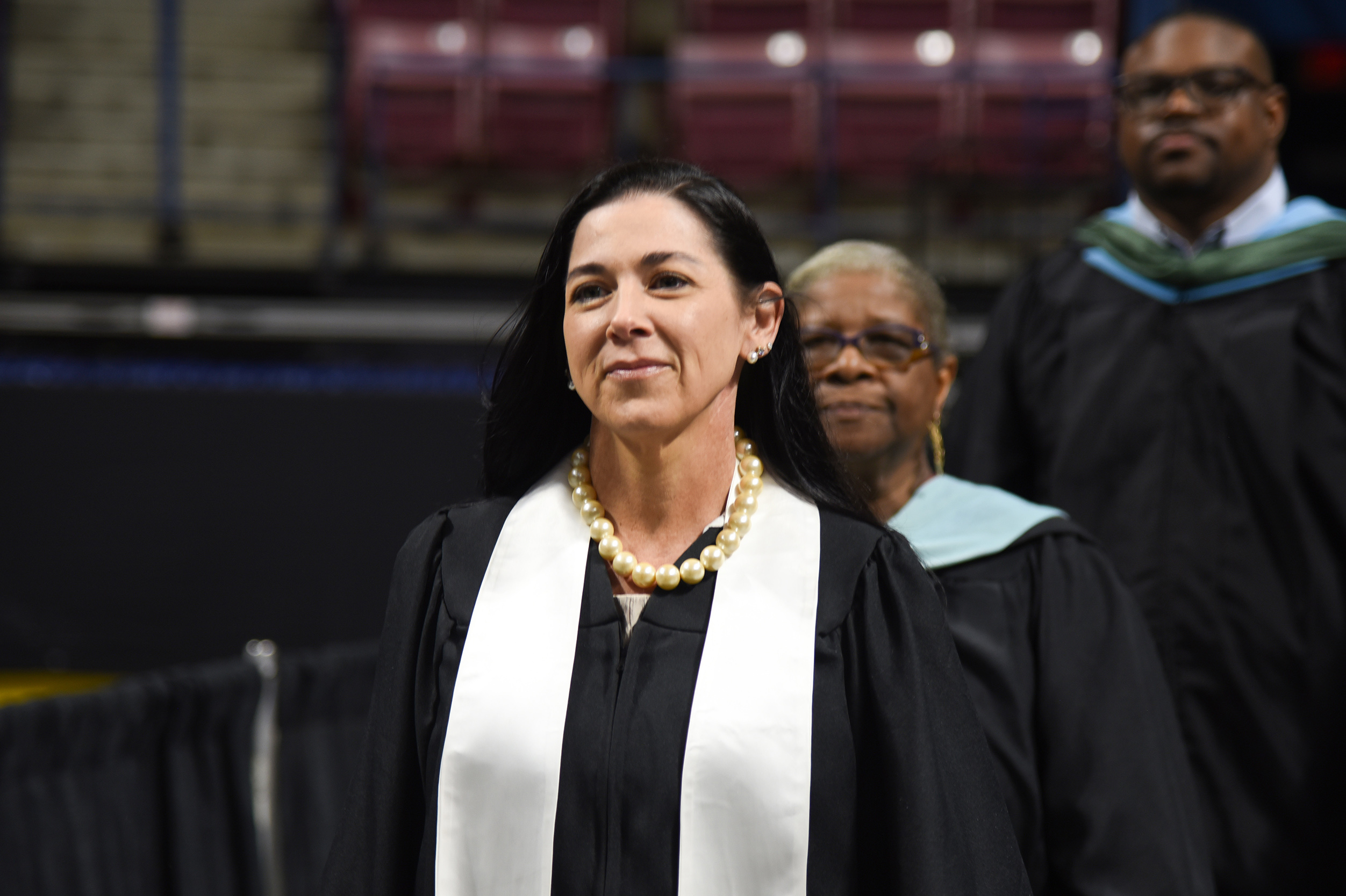 A woman in academic regalia walks in front of two others, likely at a graduation ceremony.