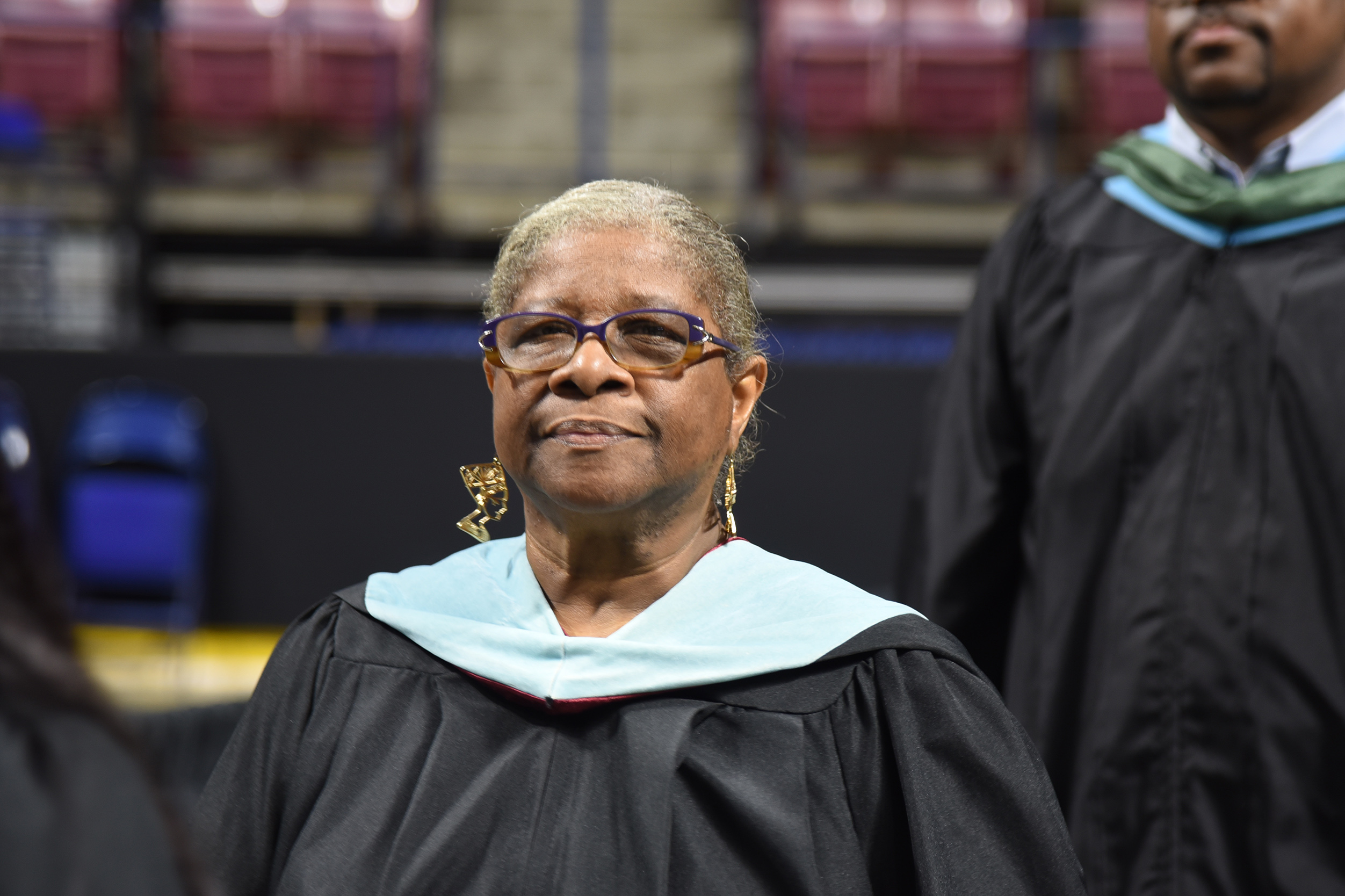 A woman in a black graduation gown with a blue stole stands in a stadium. Behind her, a person in a black gown walks.