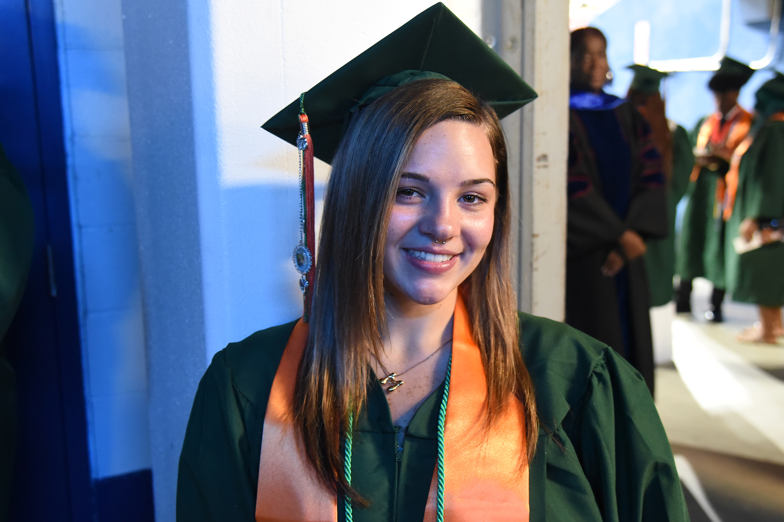 A woman in a graduation gown and cap smiles for the camera. She wears a sash and stands near others in caps and gowns.