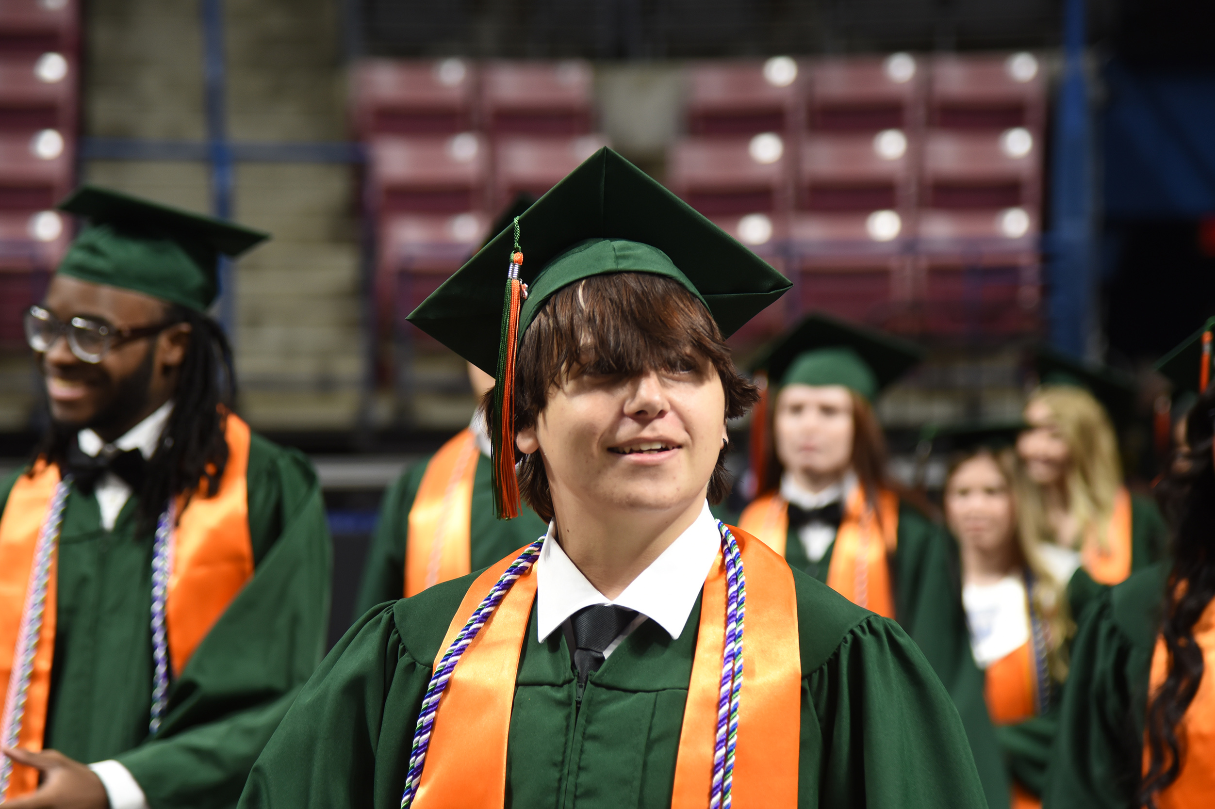 Graduates in green gowns and caps, some with glasses, stand in rows, possibly at a ceremony, with red seats in the background.