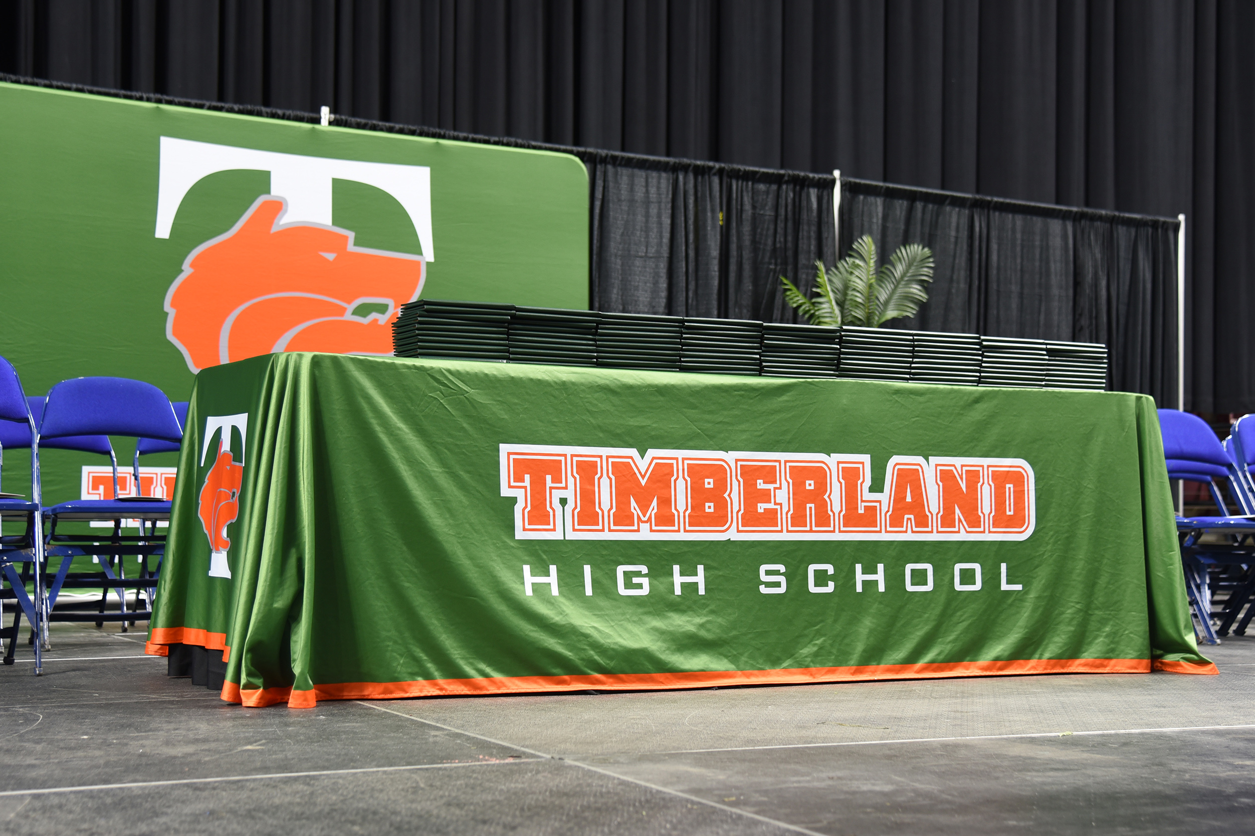 A green table with the words "TIMBERLAND HIGH SCHOOL" sits on a stage. Two blue chairs flank the table.