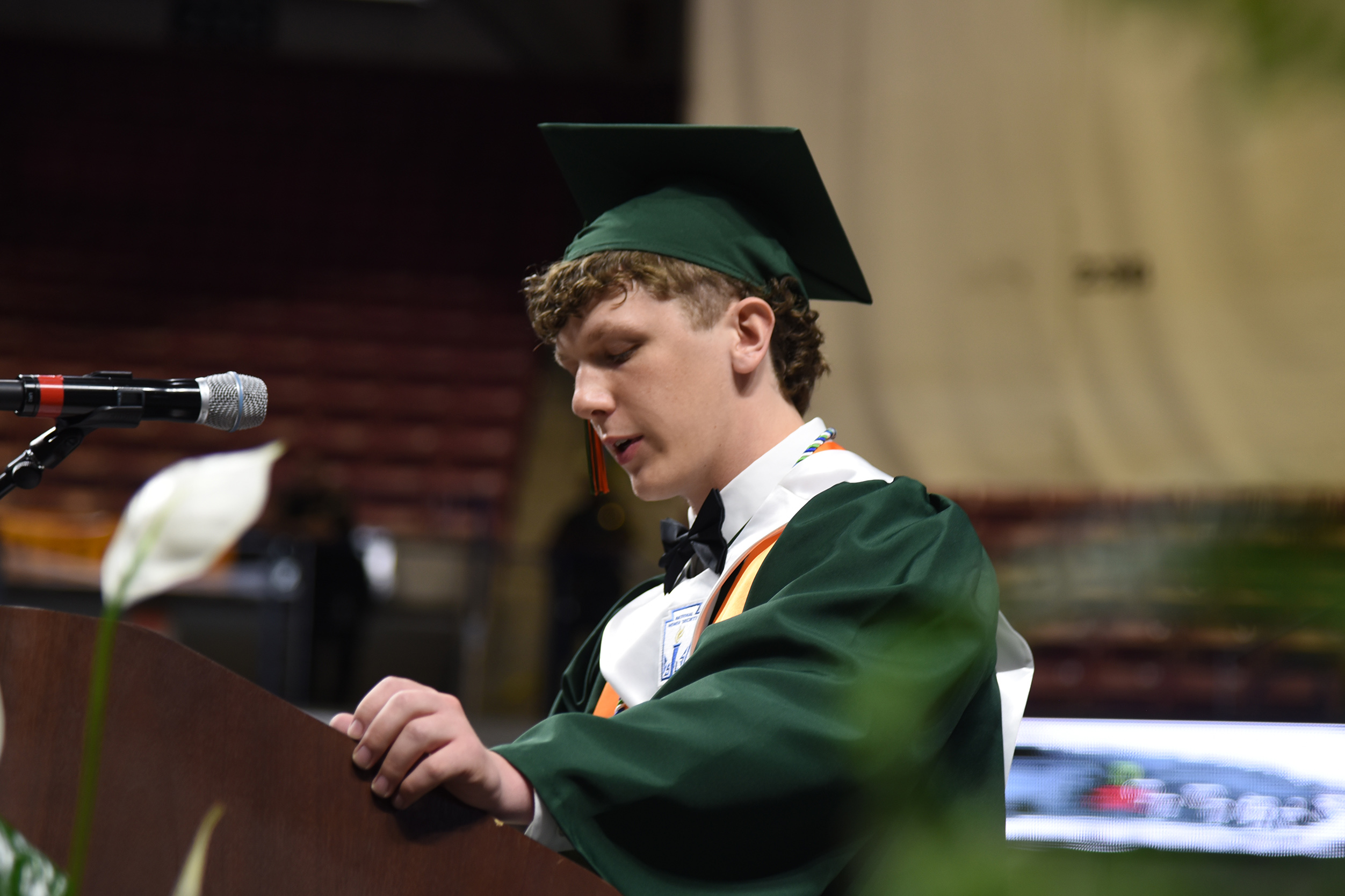 A person in a green graduation gown and cap stands at a podium with a microphone.
