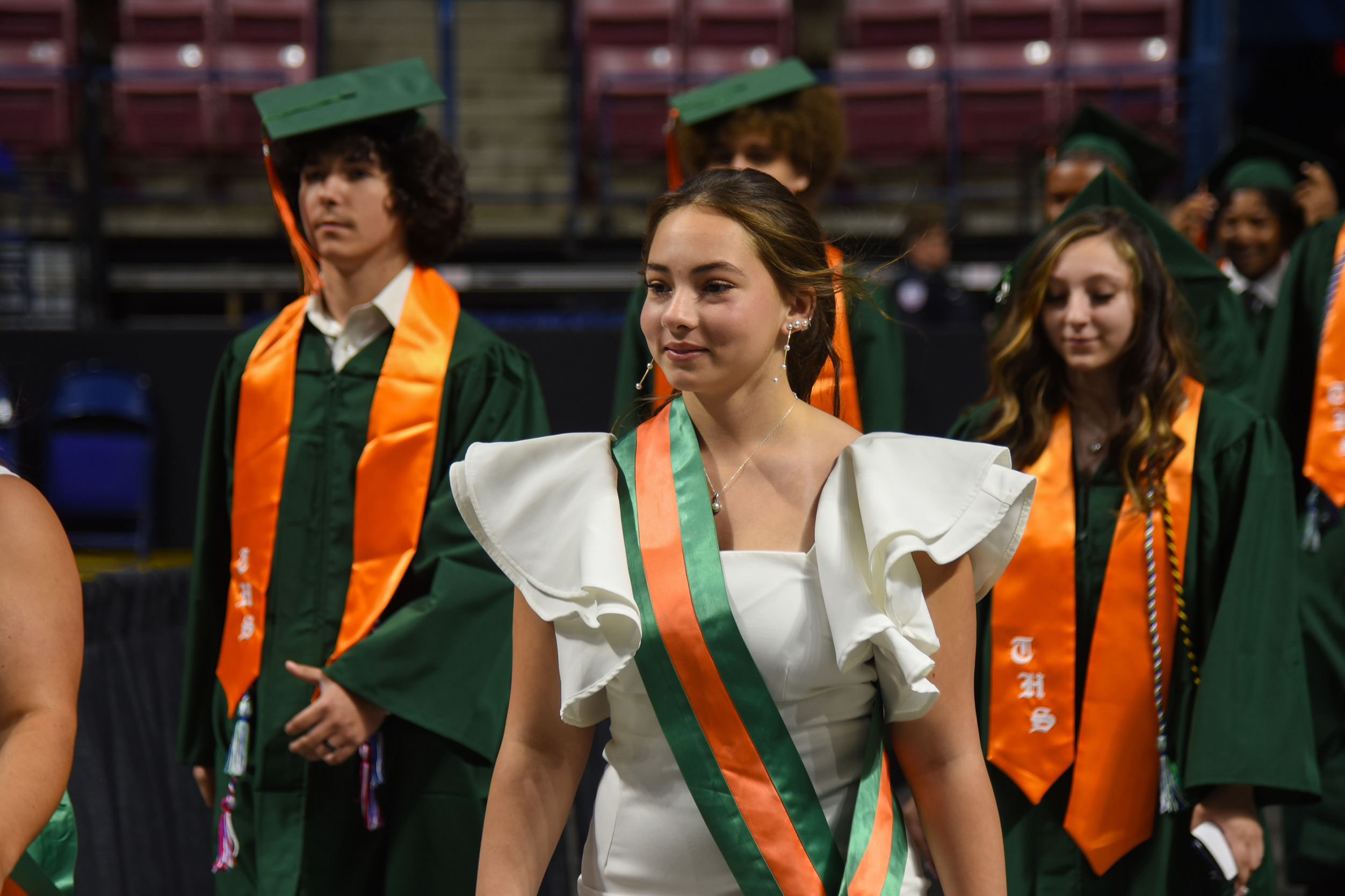 A young woman in a white gown with ruffles walks with a sash. Behind her are four other people in green and orange graduation gowns.