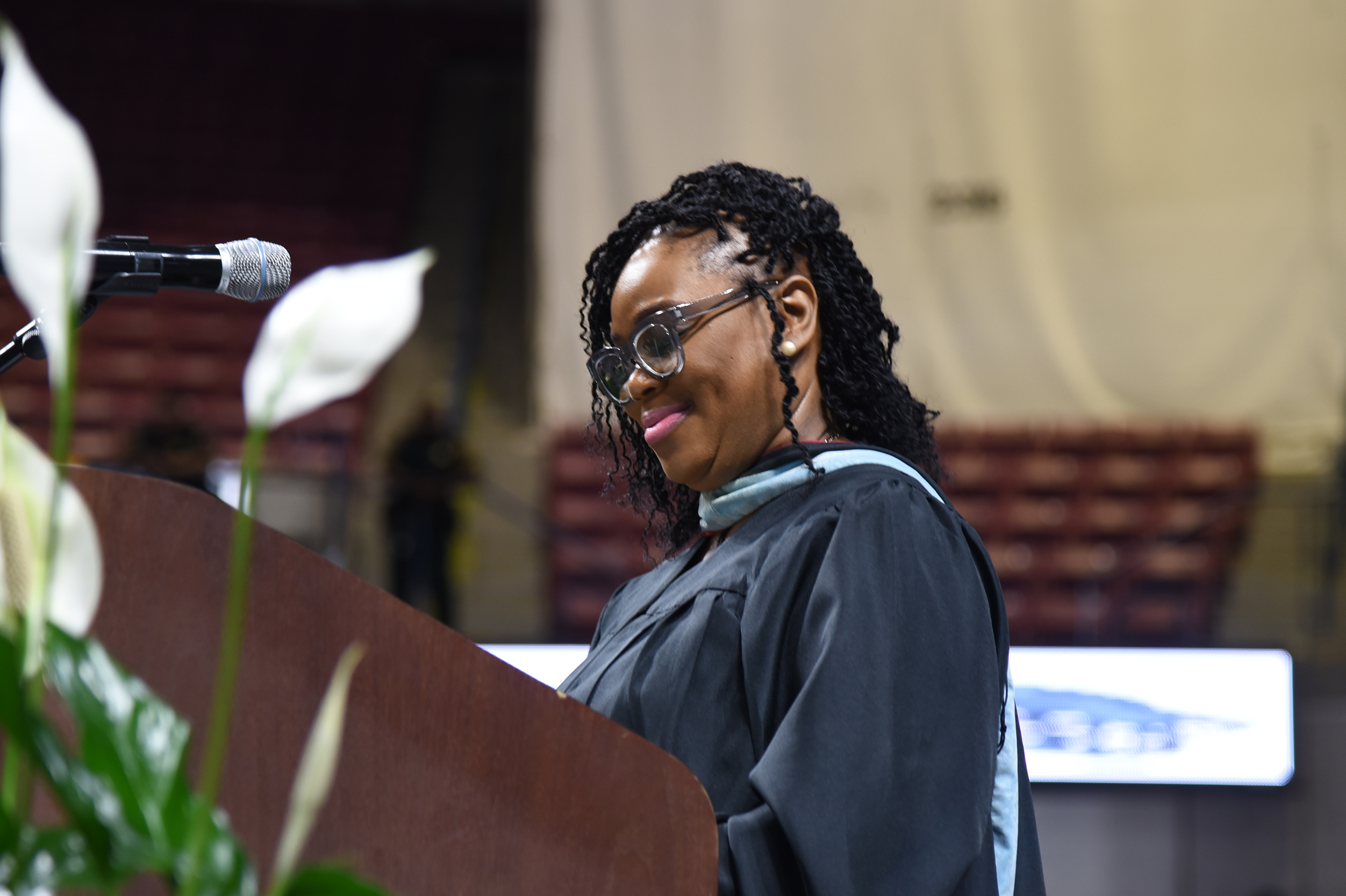 A woman in a graduation gown stands at a podium, smiling, with a microphone in front of her.