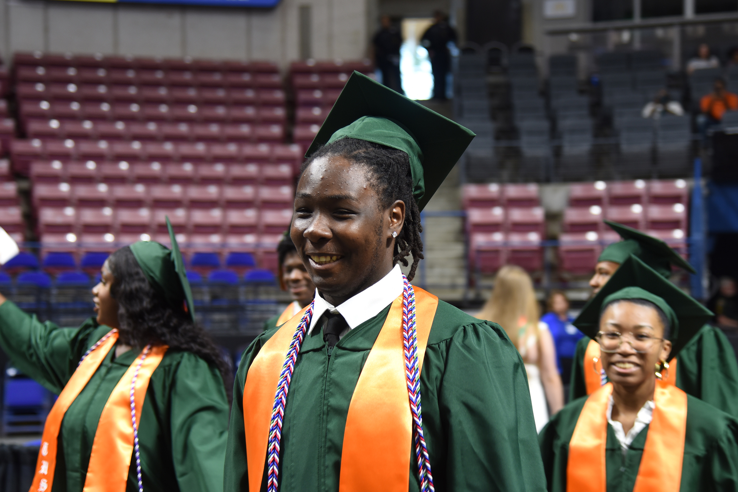 Graduates in green gowns and mortarboards walk in a line in an auditorium with red seating.