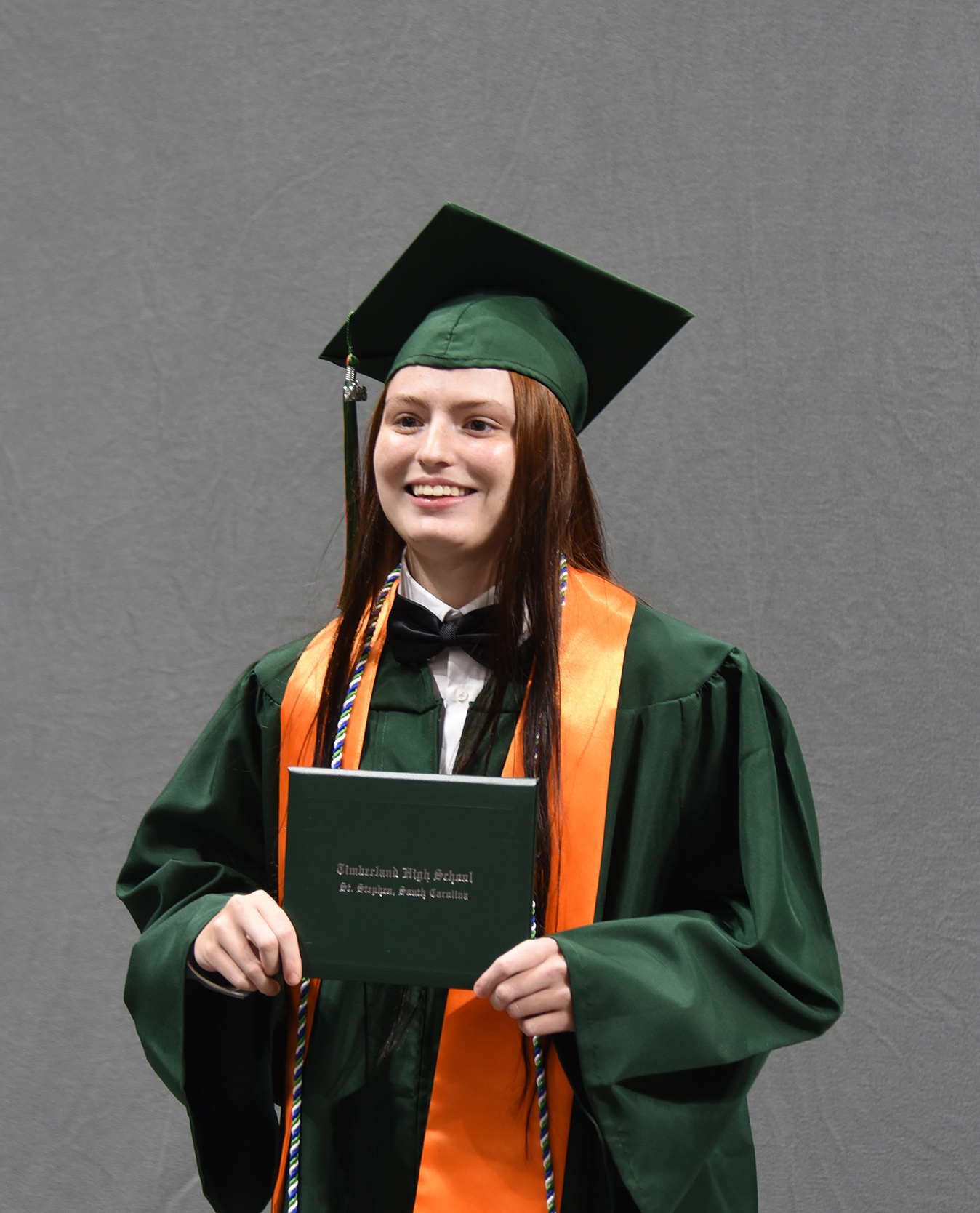 Woman in green gown and cap with tassel holds diploma. She smiles against a gray background.