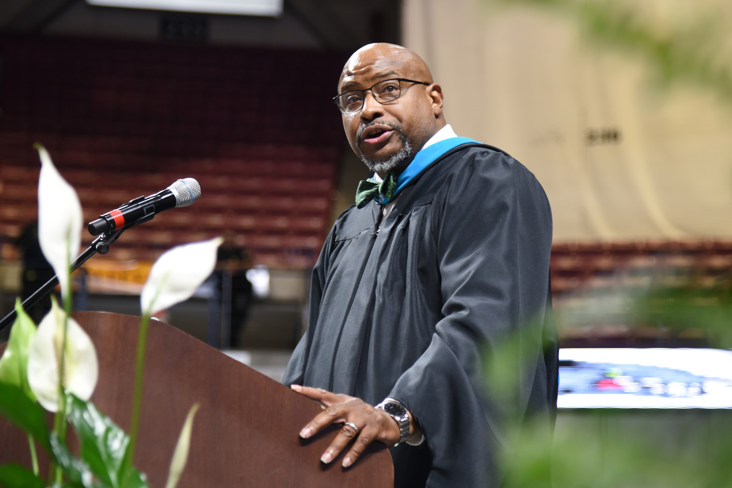 A man in a black graduation gown and bow tie stands at a podium with a microphone.
