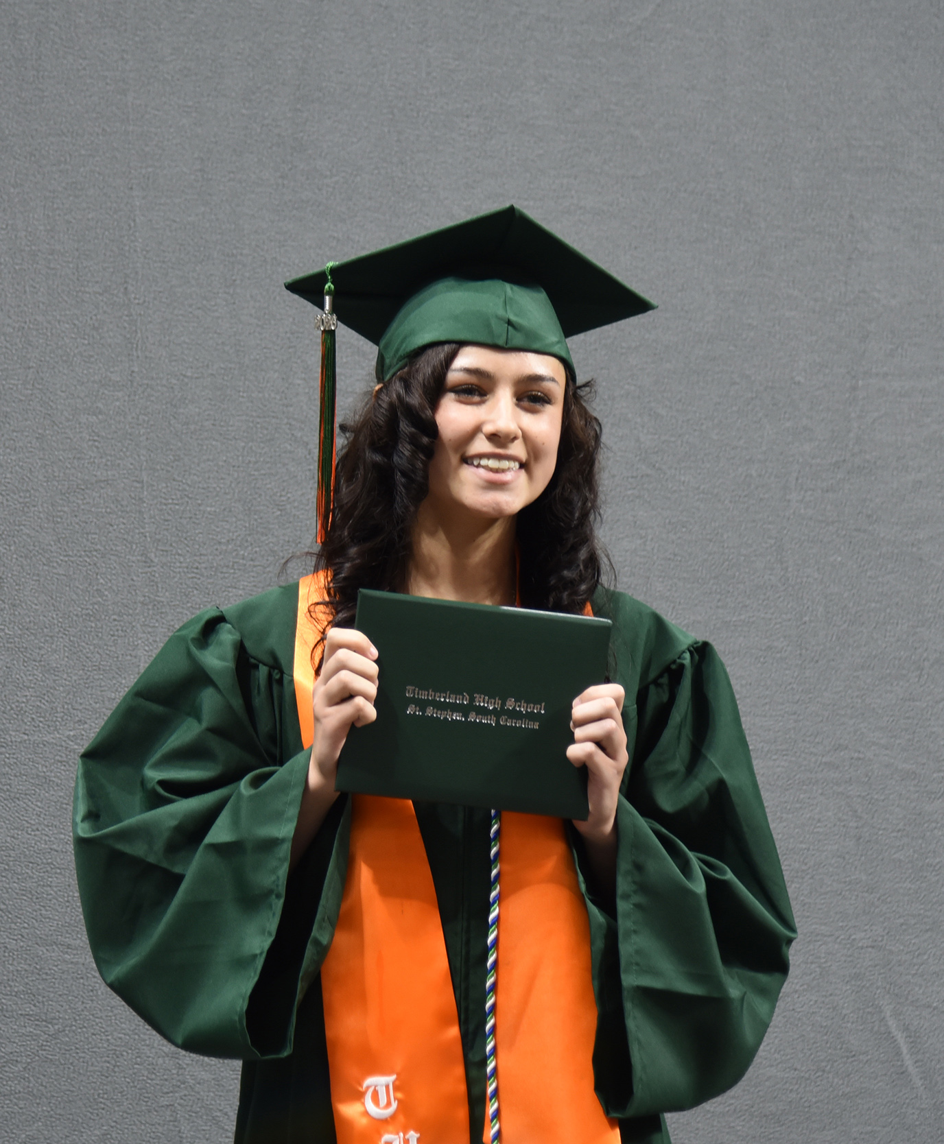 Woman in green graduation gown and cap holding diploma in front of gray background.